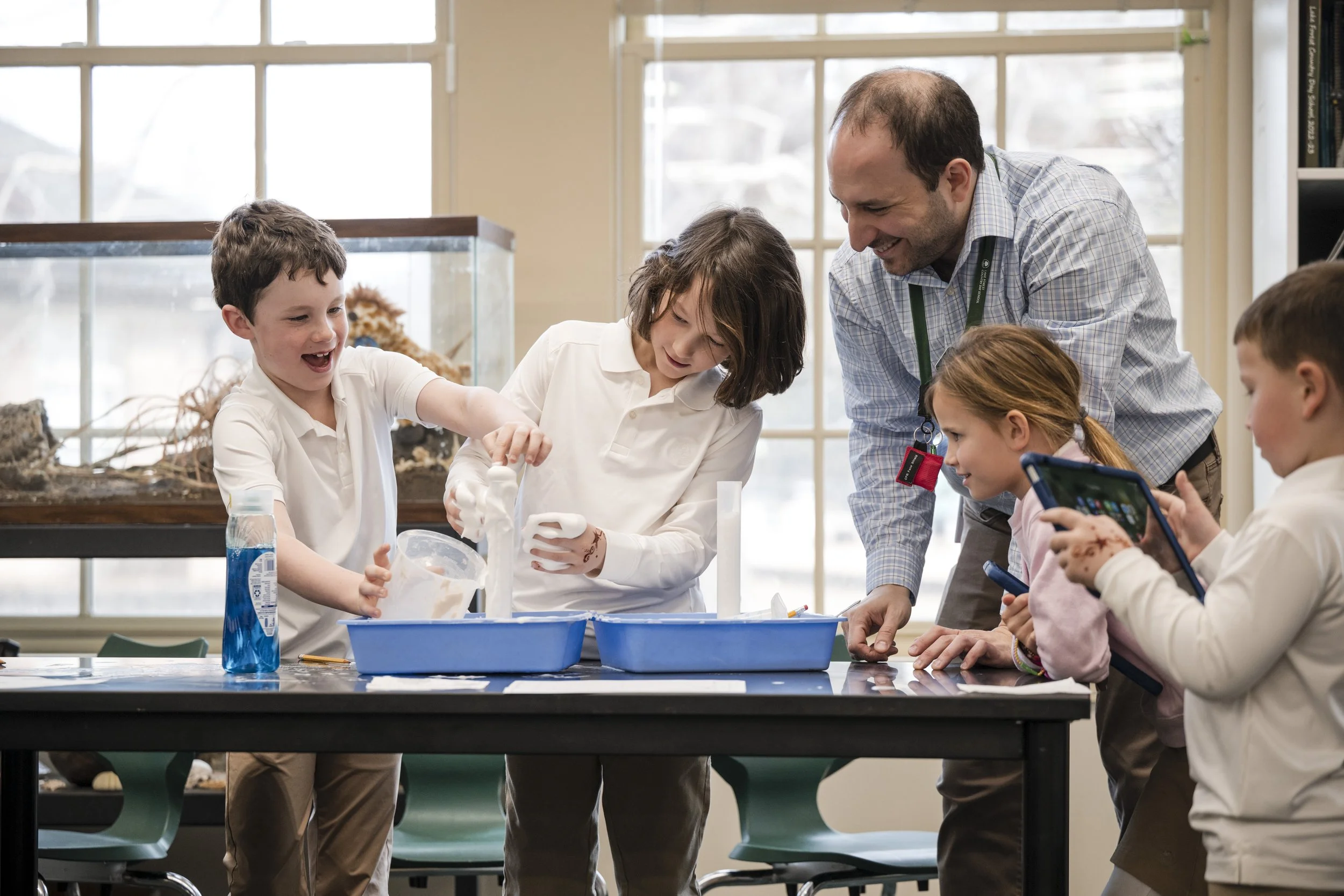 A group of children and an adult engaged in a science activity at a table in a classroom with large windows. The children appear happy and interested, with science materials and a tank with an animal in the background.