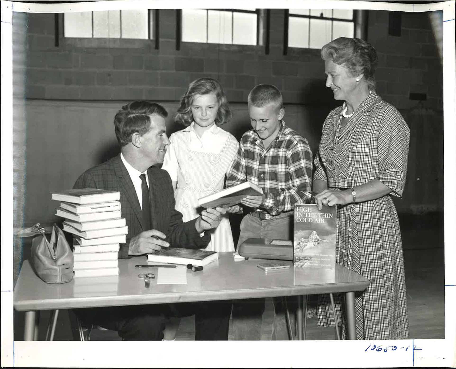 A woman with short hair and a checkered dress stands smiling at a young boy with a plaid shirt, who is holding a book, in a room with a brick wall. Two girls stand behind them, one with curly hair and the other with short hair, looking at the boy. A 