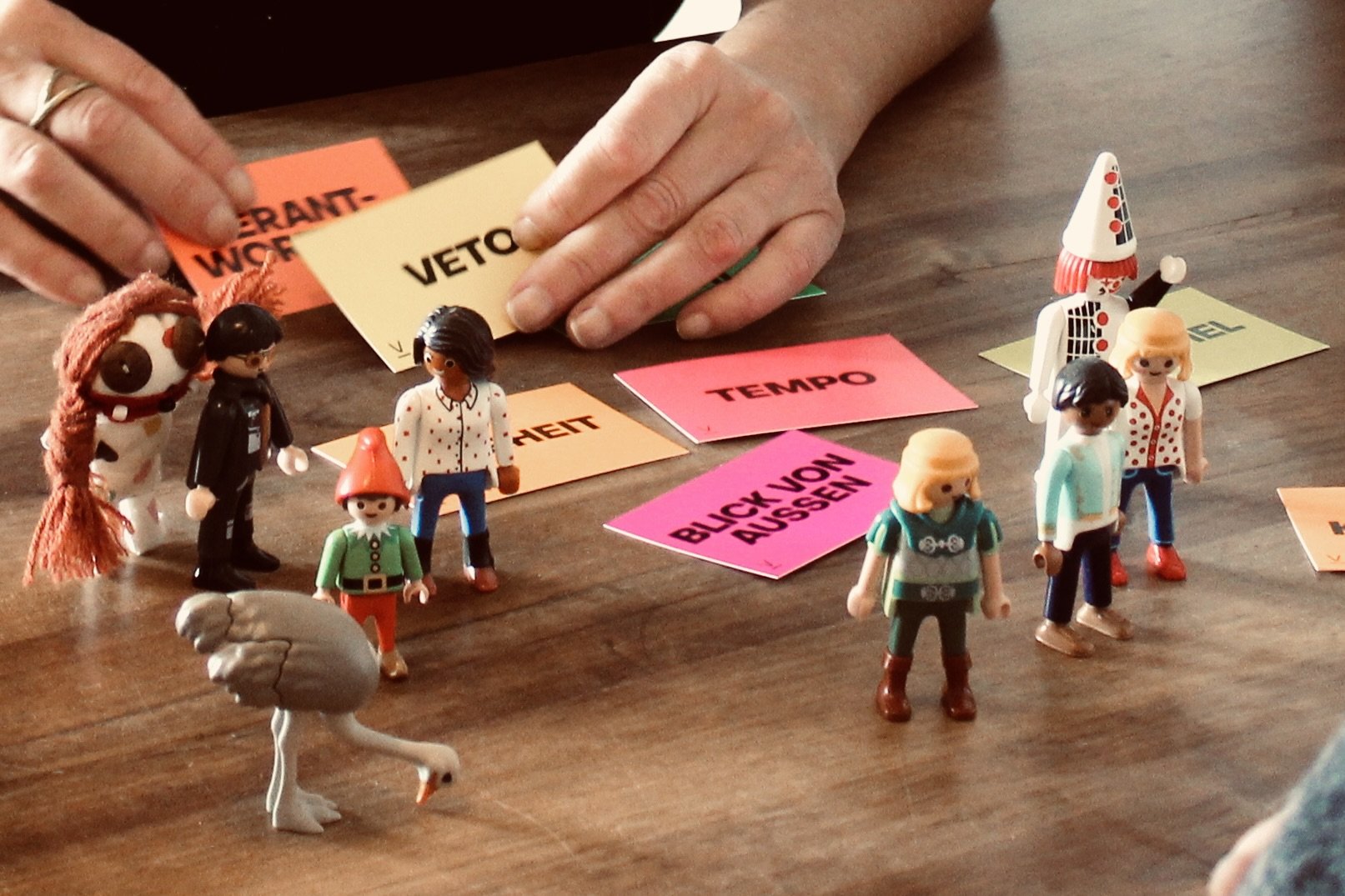 Spielzeugfiguren und bunte Karten mit deutschen Begriffen auf einem Holztisch, darunter 'VERANTWORTUNG', 'VETO', 'TEMPO', 'BLOCK-VON AUSSETZEN', um ein Lernspiel oder eine Diskussion zu symbolisieren.
