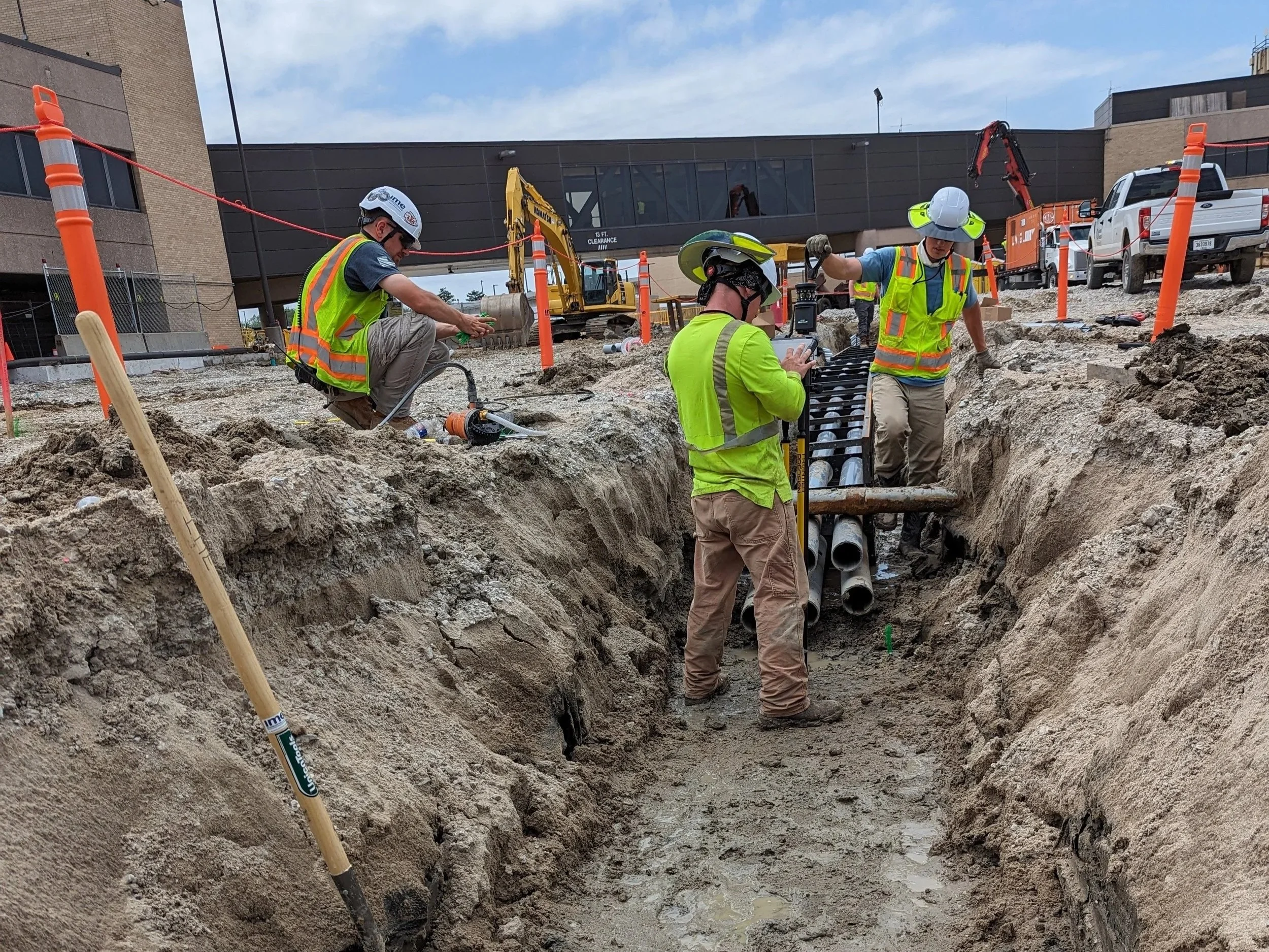 Construction workers in safety vests and helmets working on underground pipe installation at a construction site. There are orange safety barriers, construction machinery, and a building in the background.