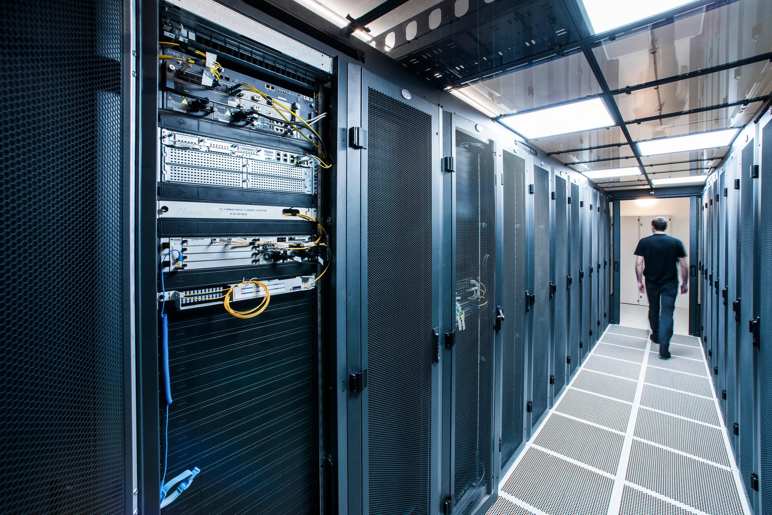 A person walking in a data center corridor with black server racks on either side, some with open doors revealing network equipment and cables. The ceiling has a grid of panels with bright lights.