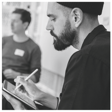 Man with a beard and beanie using a stylus and tablet, with another person in the background.