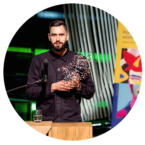 A man with a beard and dark hair standing at a podium holding a colorful molecular model, with a glass of water on the podium and colorful abstract backgrounds behind him.