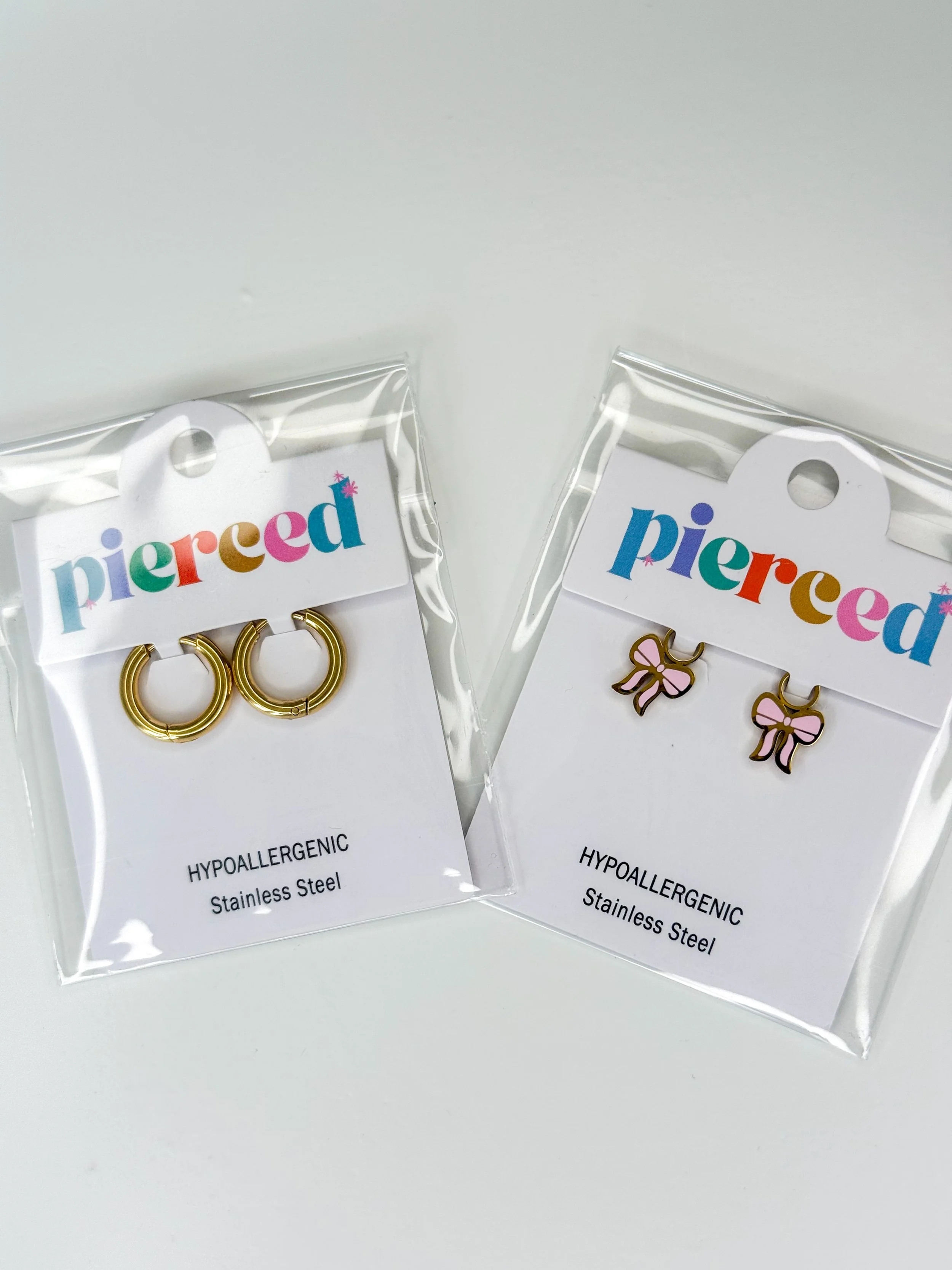 Pair of gold hoop earrings with a small pink bow charm, packaged in clear plastic with a white label that reads "pierced" in colorful letters and "hypoallergenic stainless steel".