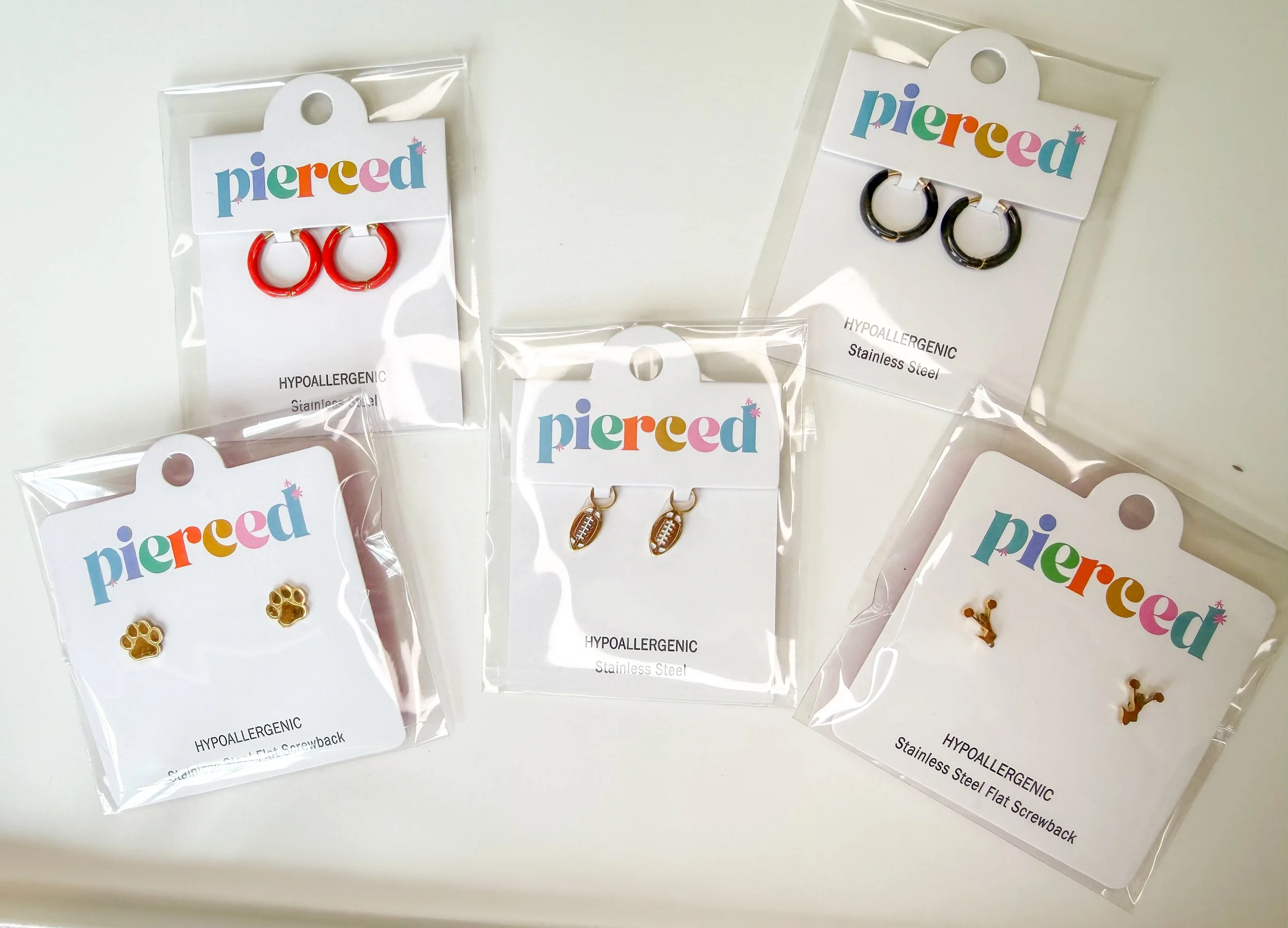 Several pairs of stainless steel earrings, including red hoops, black hoops, gold animal paw, and football-shaped earrings, each on individual cards with the brand name 'Pierced' in colorful letters.