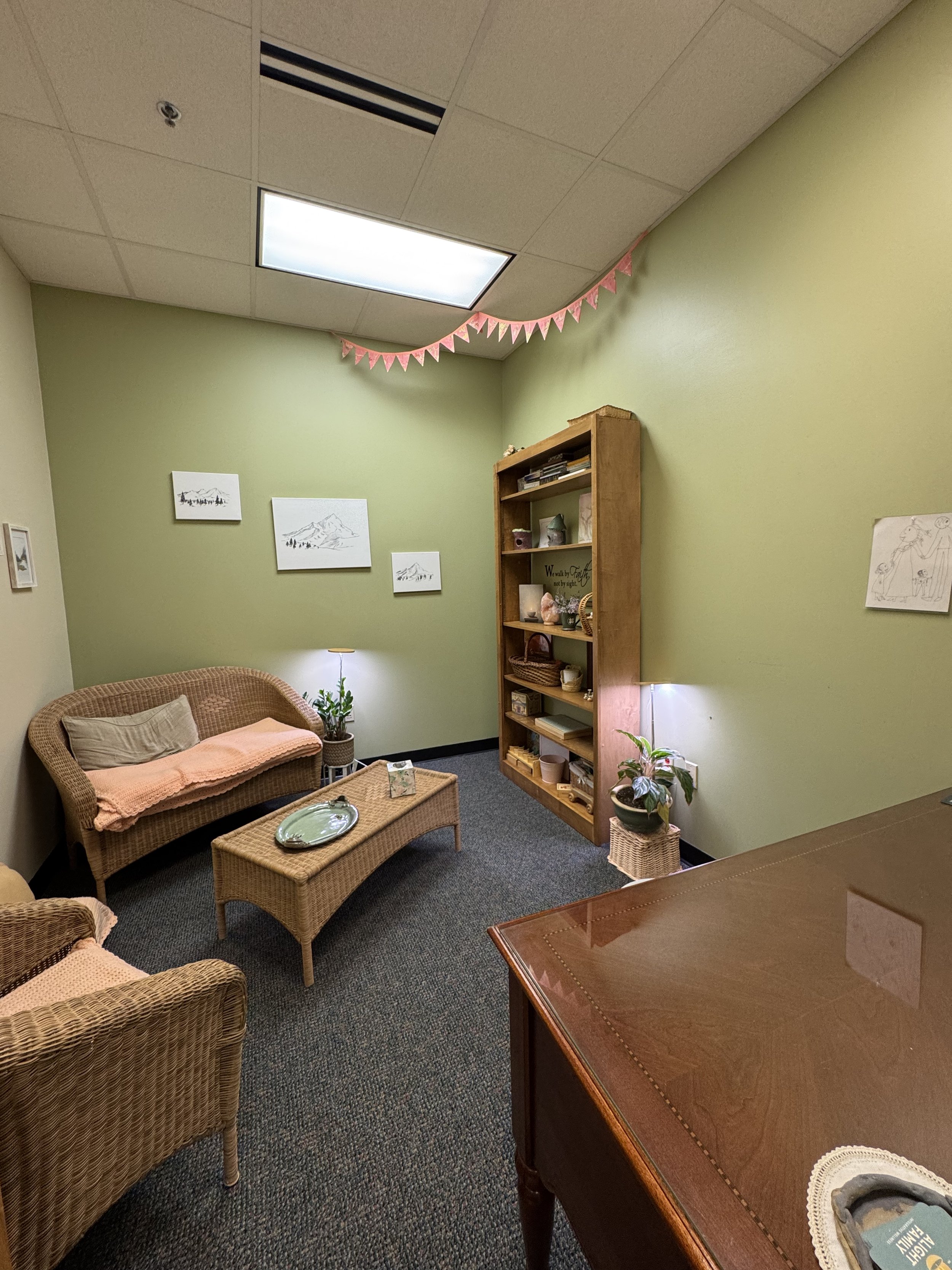 A cozy room with green walls and framed artwork, featuring wicker furniture including a loveseat and coffee table, a wooden bookshelf with decorative items, and a potted plant, with pink triangular banners hanging from the ceiling and light fixtures providing soft lighting.
