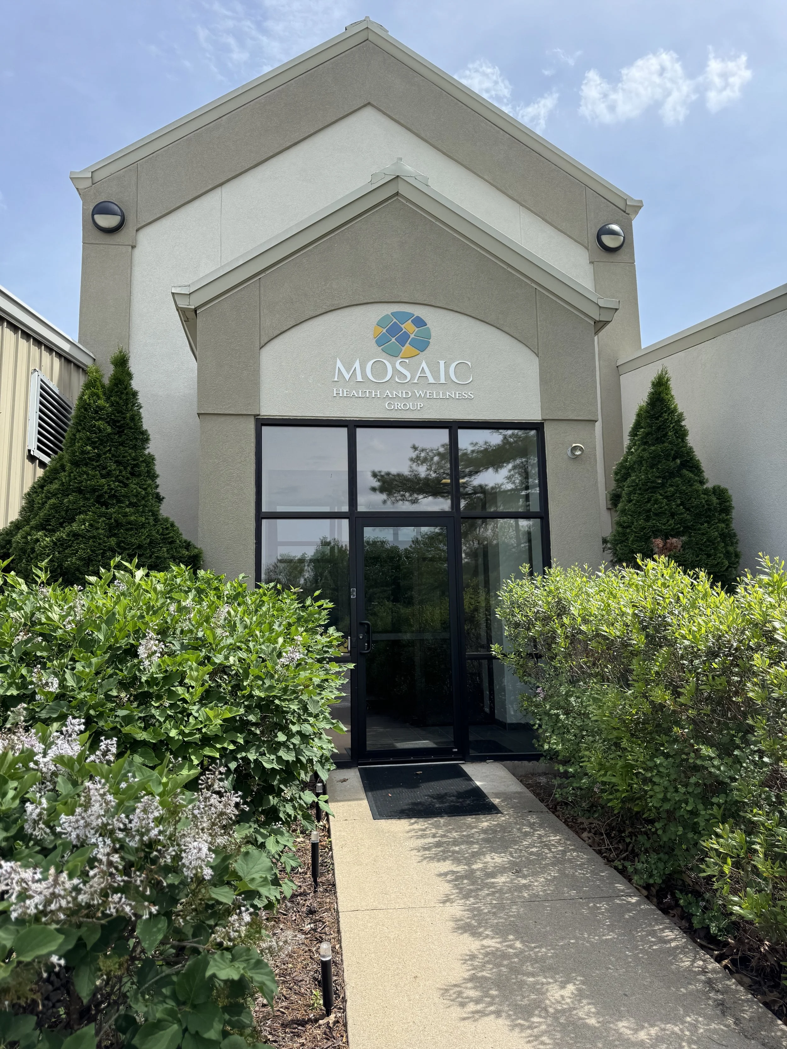 Building entrance with signage reading 'MOSAIC Health and Wellness Group,' surrounded by green bushes, trees, and a concrete pathway leading to the door.