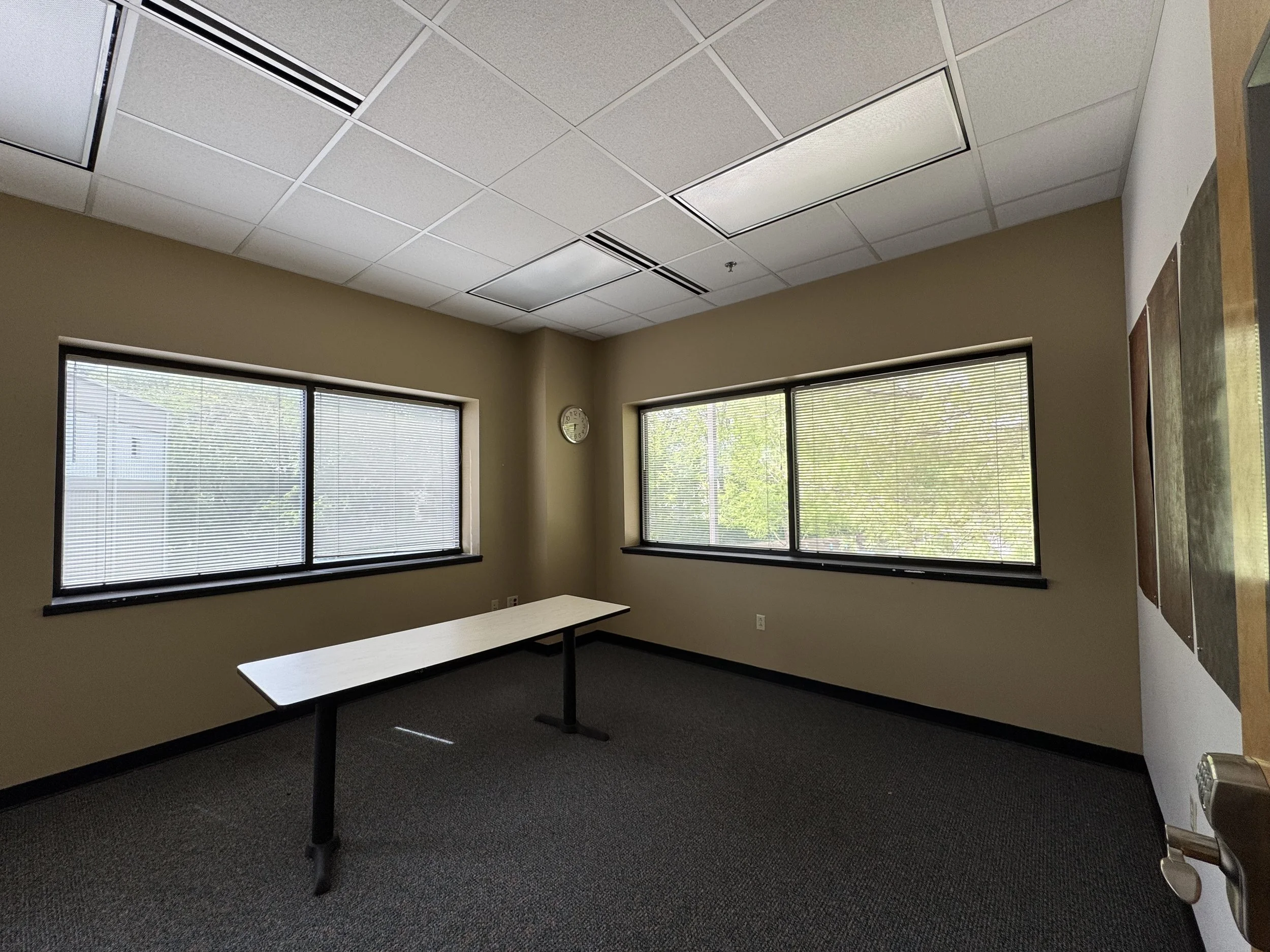 An empty office room with beige walls, two large windows with blinds, a clock on the wall, a long table, and ceiling tiles with fluorescent lights.