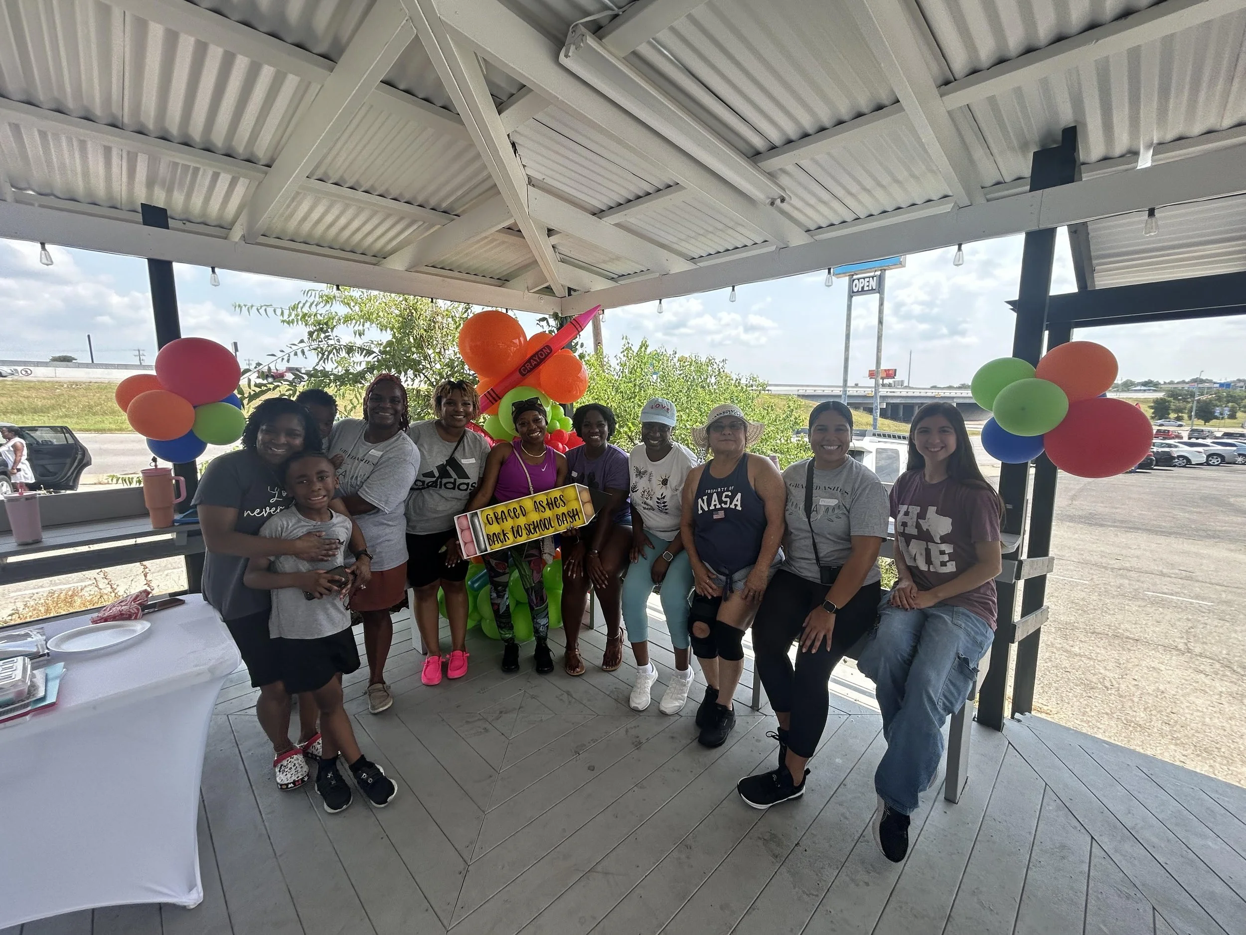 A group of smiling people, including children and adults, standing together under an outdoor covered area decorated with colorful balloons and a sign that reads "GRACED ASKS BACK TO SCHOOL BASH." In the background, there is a parking lot, trees, and a highway.