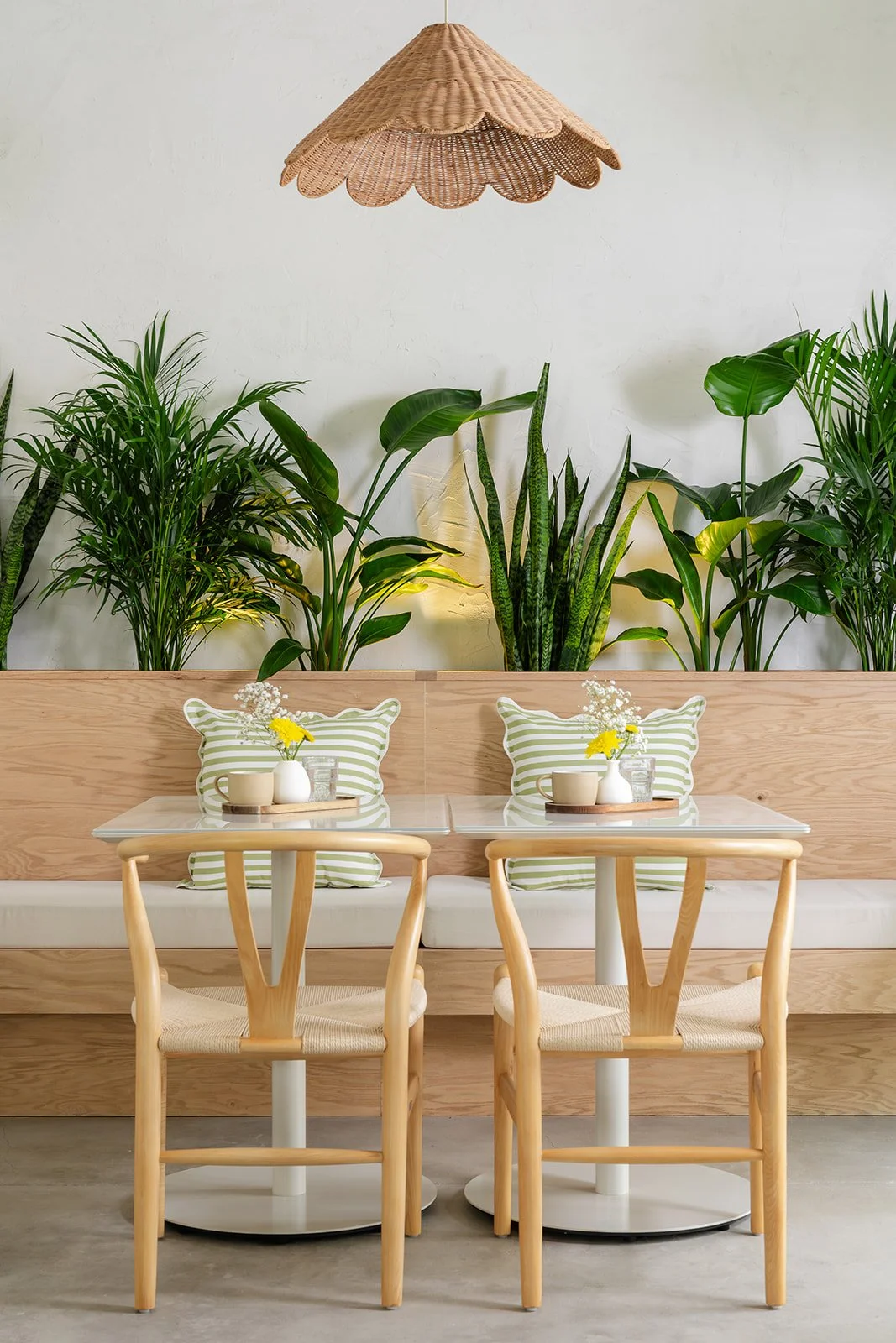 A cozy indoor dining area with two small tables, each with a striped pillow, small vase with yellow and white flowers, and a glass of water. Behind the tables is a wooden bench with green leafy plants and a hanging wicker lampshade overhead.