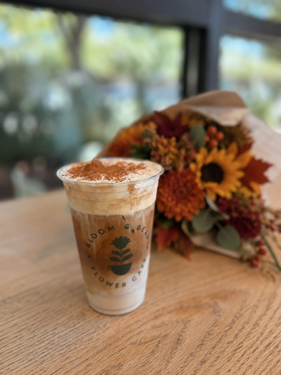 Iced coffee with whipped cream and cocoa powder on top, placed on a wooden table with a bouquet of fall flowers in the background.