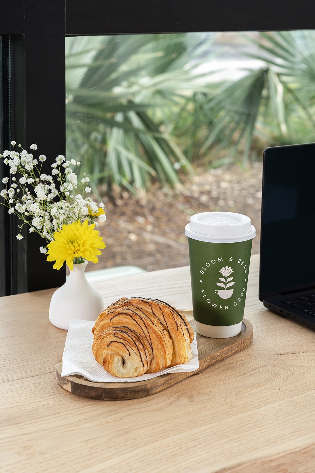 Pastry with chocolate drizzle on a wooden table, next to a green paper coffee cup with a white lid, a small white flowers and a yellow flower in a white vase, and a black laptop, with greenery outside the window.