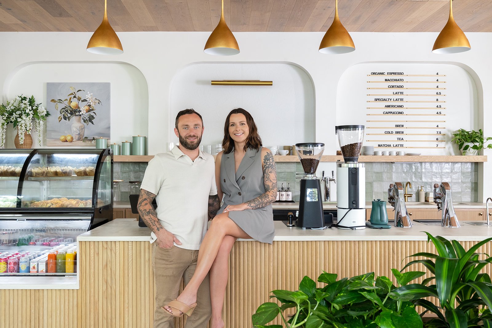 A man and a woman smiling behind the counter of a modern coffee shop. The woman is sitting on the counter with her legs crossed, and the man is standing next to her with his hand in his pocket. The background features a menu board, coffee equipment, and decorative plants.