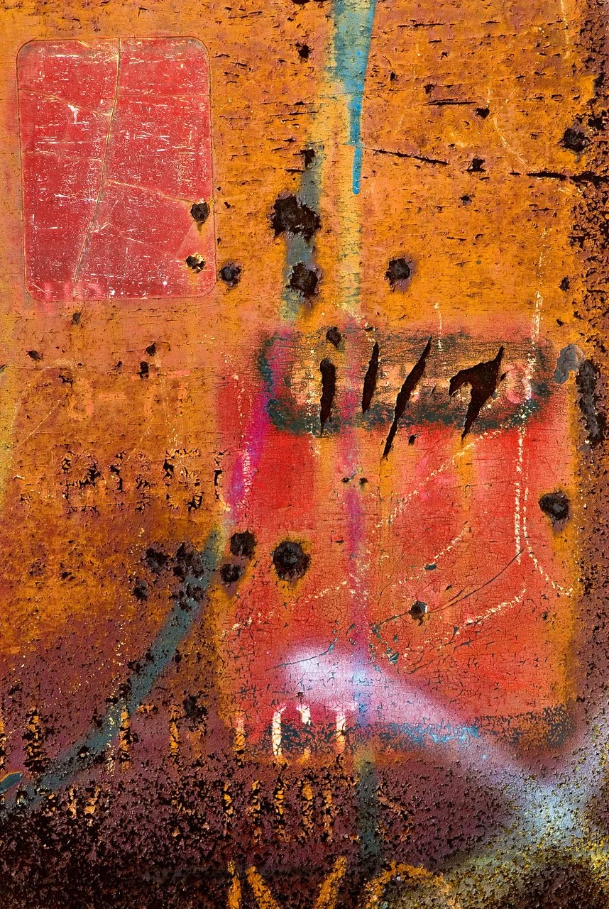 Close-up of a rusty, weathered metal surface with peeling paint, scorch marks, and graffiti in red, black, and hints of pink and blue.