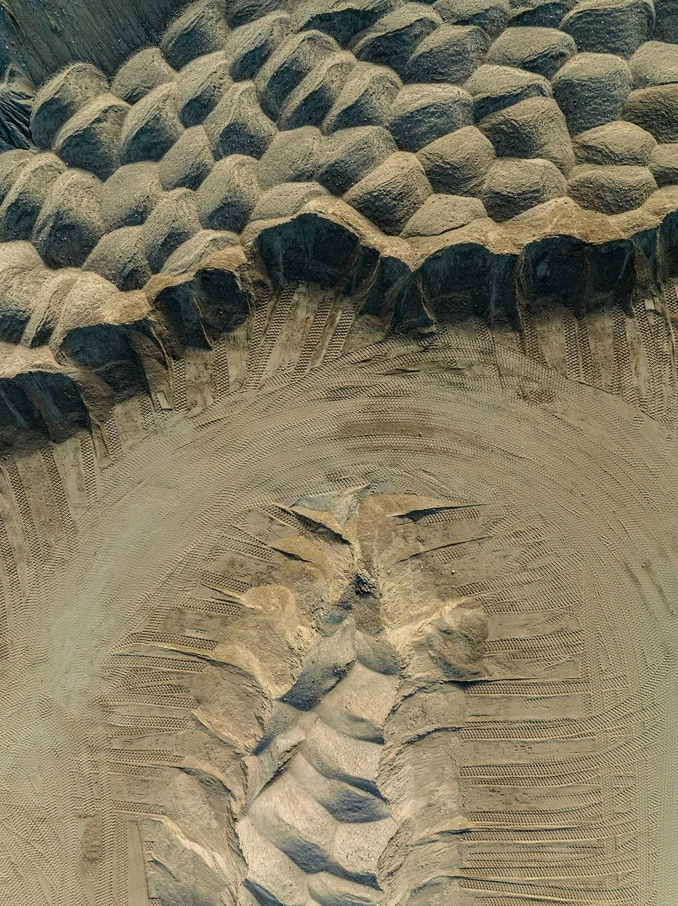 Close-up view of a large tire tread pattern, showing detailed grooves and tire marks in the dirt, likely from a construction vehicle or heavy machinery.