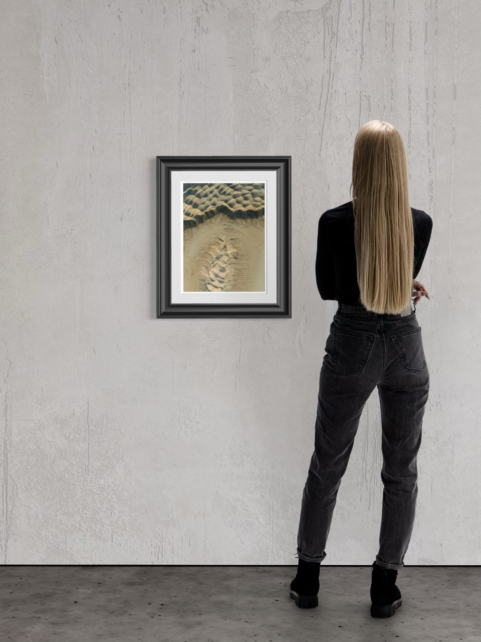 A woman with long blonde hair wearing a black jacket and black jeans stands in front of a framed artwork on a plain gray wall, viewing the art."}
