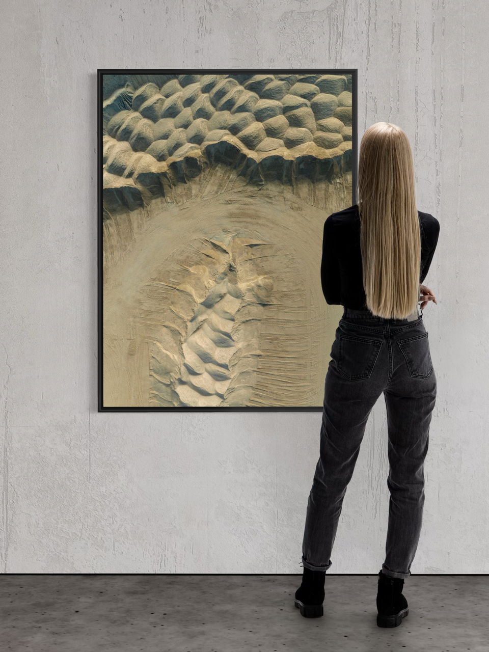 A woman with long blonde hair, wearing black jeans and a black top, stands in front of abstract textured artwork on a white gallery wall.