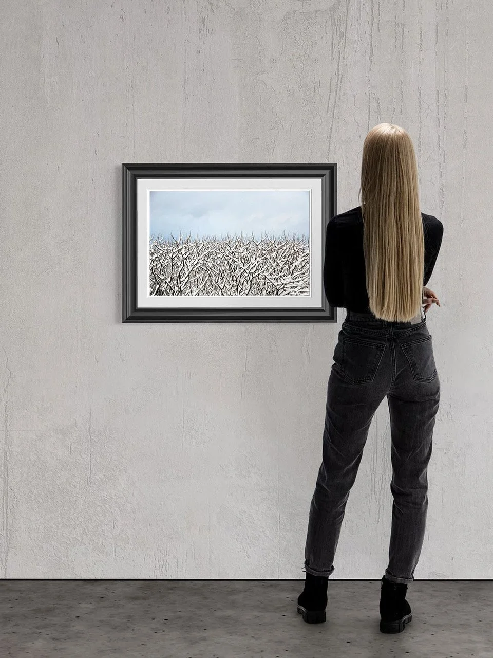 A woman with long blonde hair, dressed in black jeans and a black jacket, stands facing a framed winter landscape painting of snow-covered trees on a light gray wall in an art gallery.