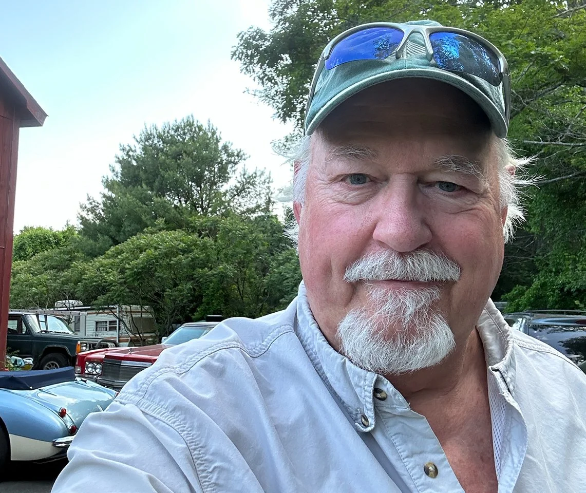 A smiling older man with a white beard and mustache wearing a light-colored button-up shirt, a baseball cap, and sunglasses on his cap. He is outdoors with green trees and parked cars in the background.
