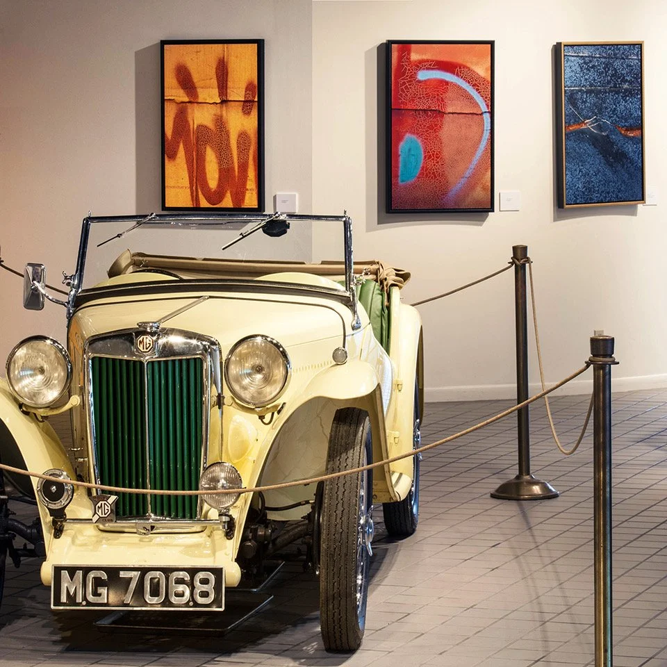 A vintage cream-colored MG convertible car displayed in an art gallery with framed abstract paintings on the wall behind it, surrounded by a rope barrier.
