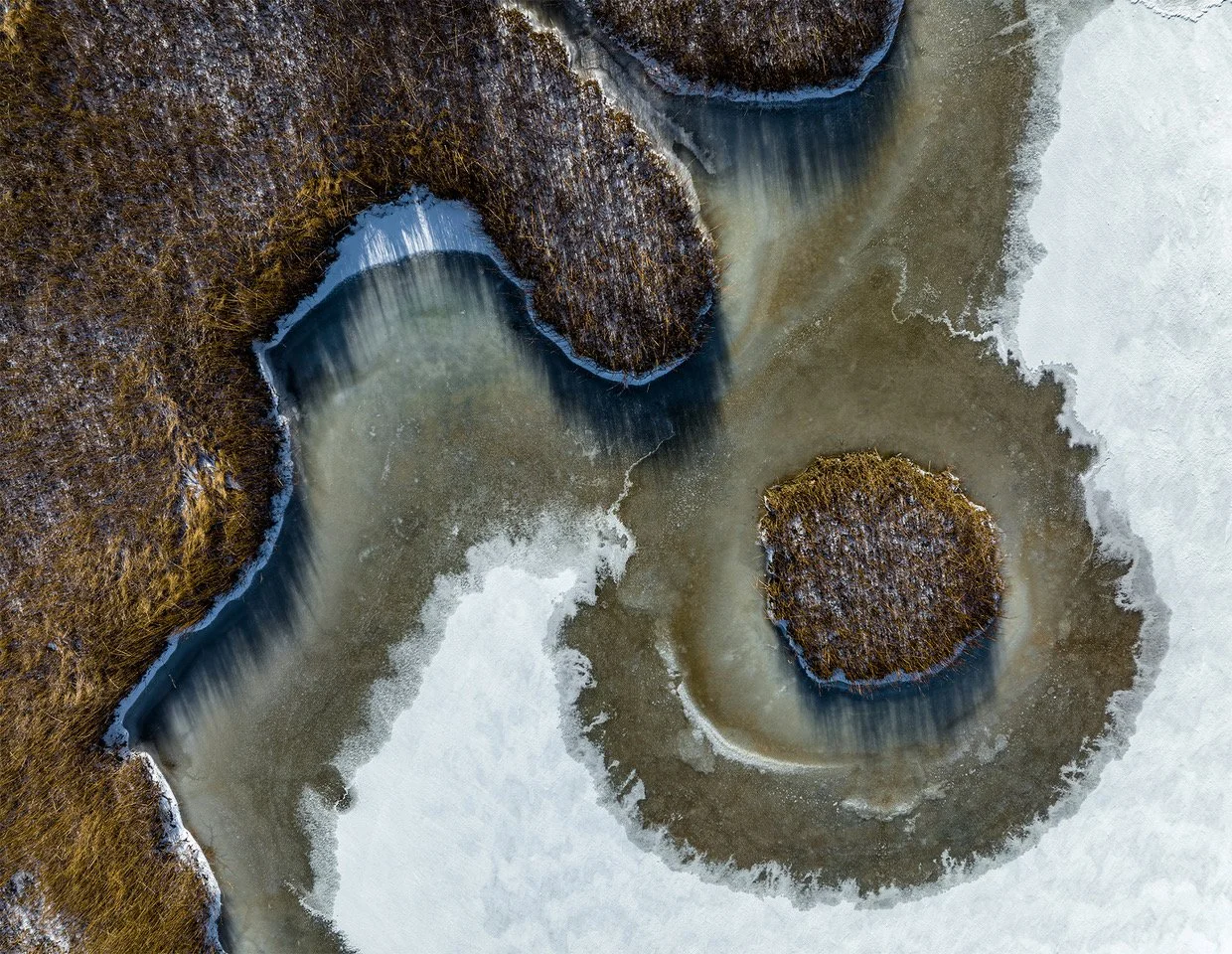 Aerial view of a river flowing around a small island covered in dry brown grass, with icy edges along the water.