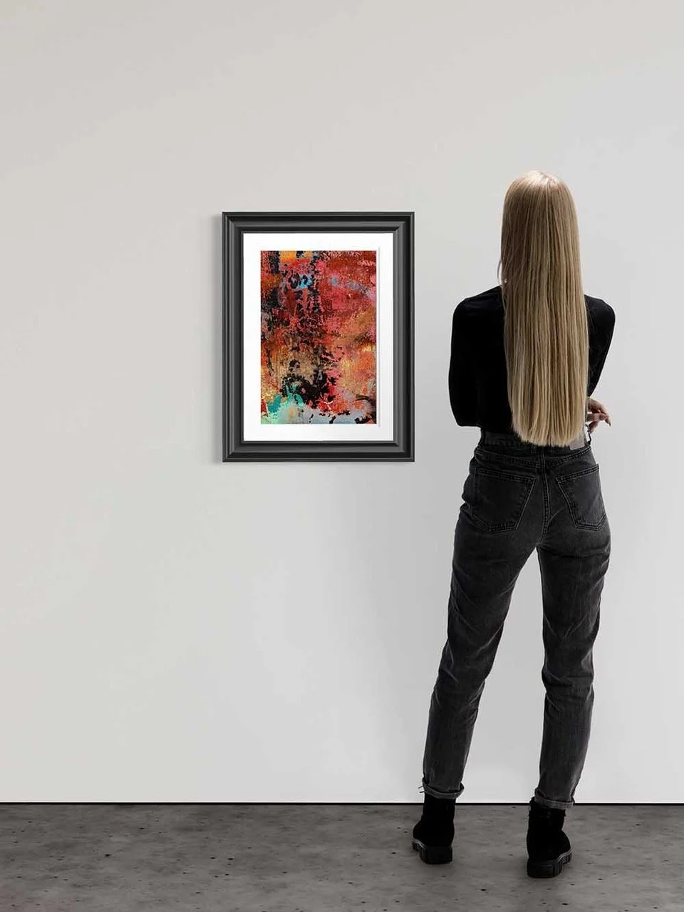 A woman with long blonde hair observing an abstract painting in a black frame on a white gallery wall.