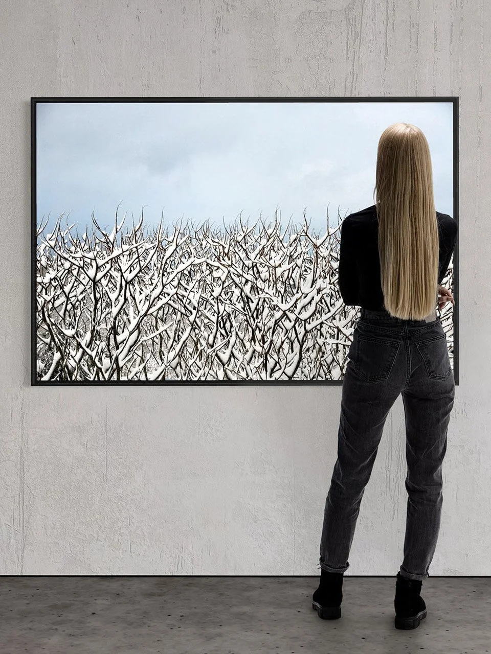 A woman with long blonde hair, dressed in black, is standing in an art gallery, observing a large framed picture of snow-covered tree branches against a cloudy sky.