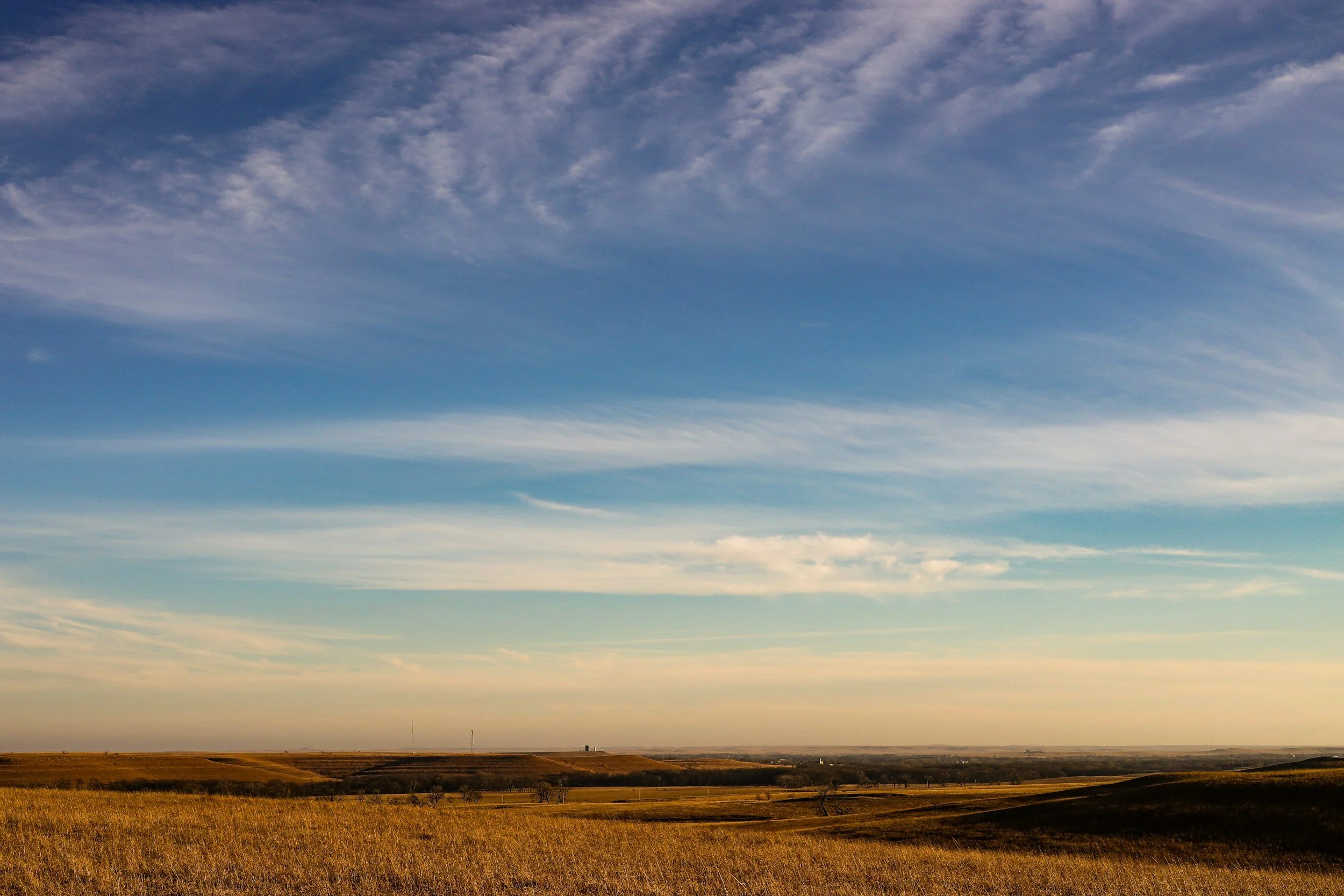 A wide landscape view of flat, open fields with a small distant horizon, under a vast sky with scattered clouds in shades of blue and white.