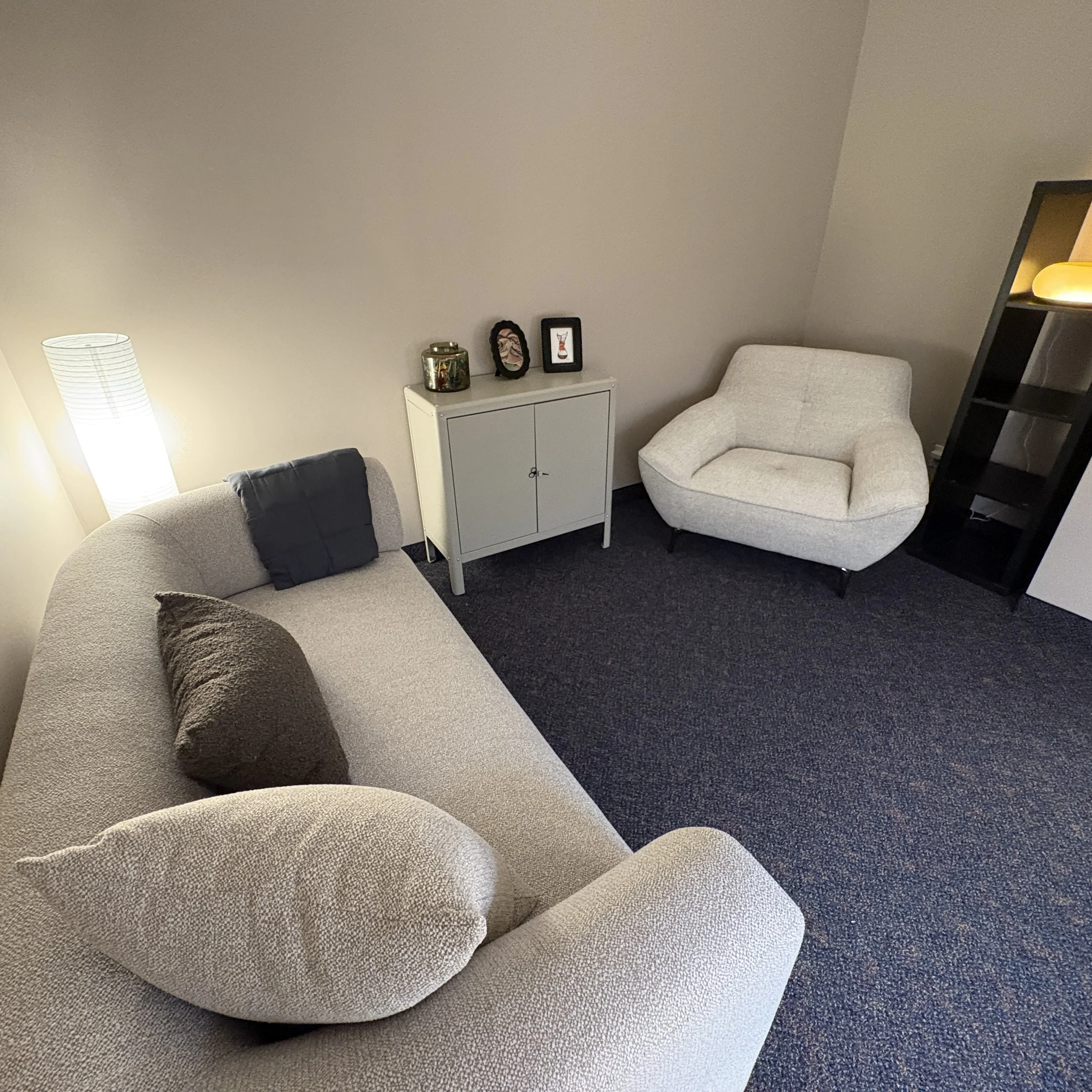 Living room corner with two beige upholstered chairs, a black cushion, a white cabinet, framed artwork, a small decorative jar, and a white floor lamp, on dark carpet.