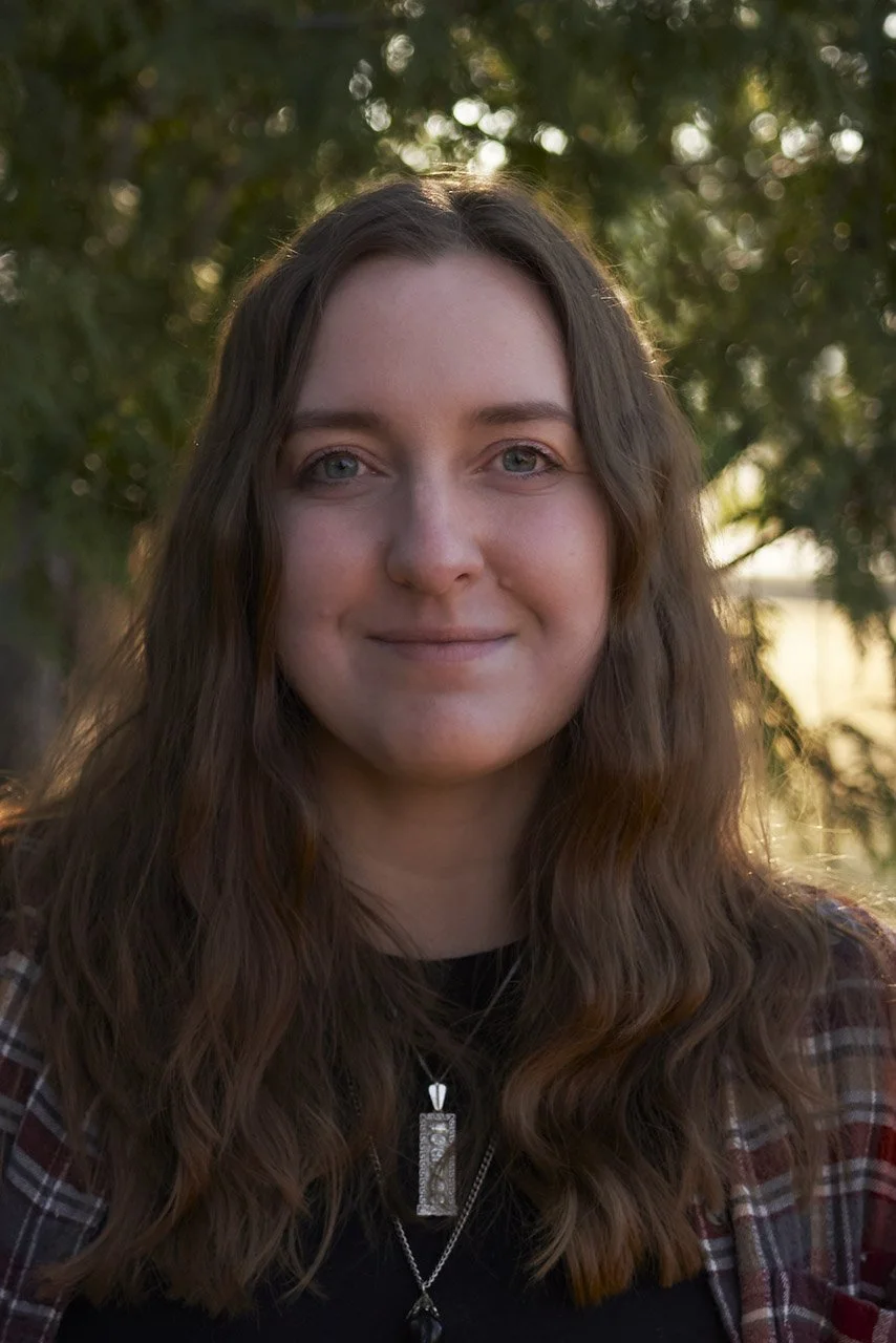 A young woman with long, wavy brown hair and blue eyes standing outdoors in front of green trees at sunset.