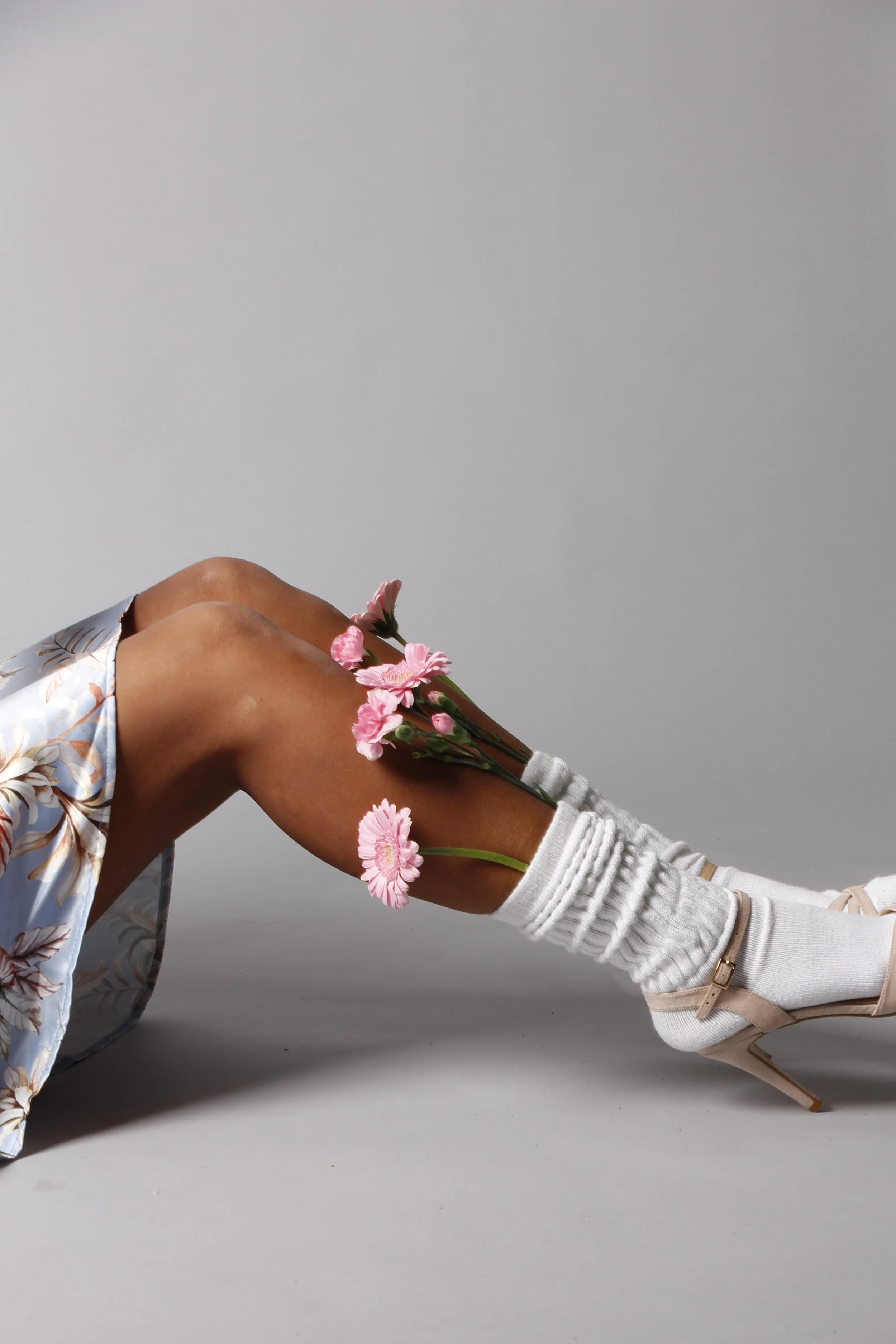 Ein Mensch liegt auf der Seite, trägt weiße Socken mit Rippmuster, eine weiße Hose und eine Bluse mit Blumenmuster. Seine Armbeuge ruht auf der Unterlage, und auf seinem Arm liegen pinke Blumen.