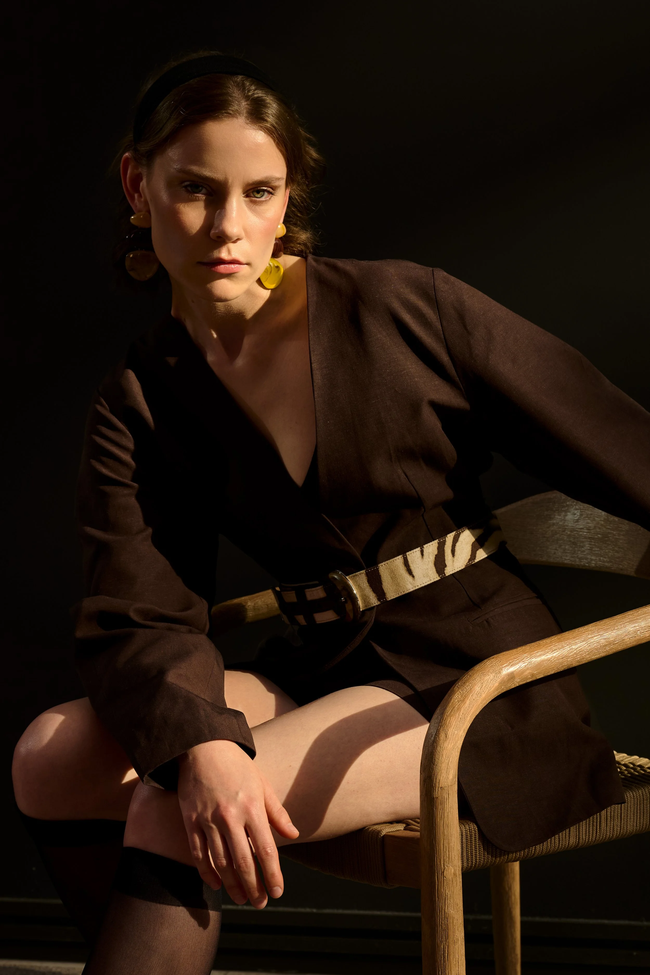 Ein Model sitzt an einem Holzstuhl, trägt dunkle Kleidung und hat lange, dunkle Haare mit einem schwarzen Haarband. Der Hintergrund ist dunkel.