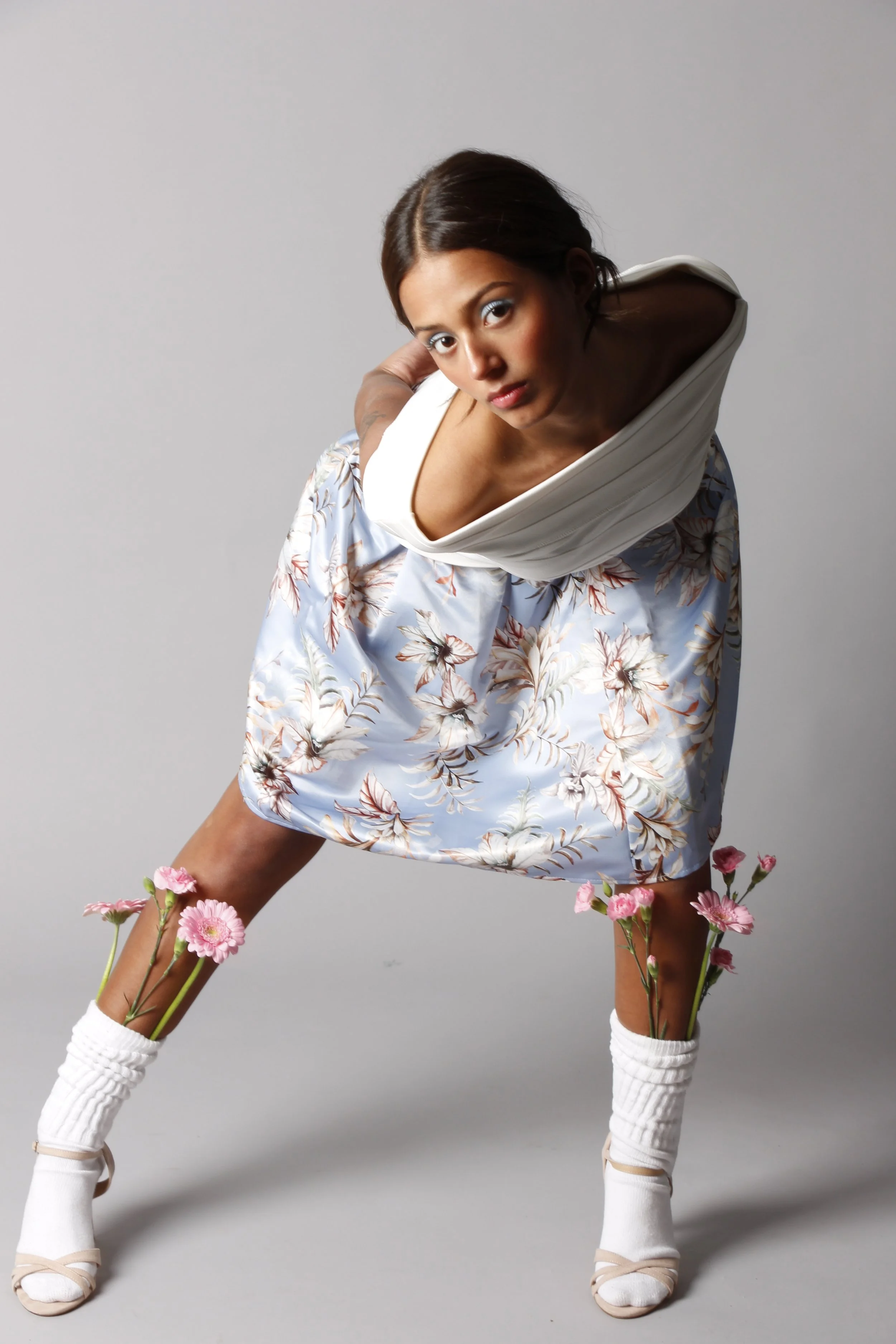 Eine junge Frau mit dunkelbraunen Haaren in einem weißen Kleid mit Blumenmuster. Sie steht in einer ungewöhnlichen Haltung, bei der ihre Beine von Blumen durchstossen sind und hält den Oberkörper leicht nach vorne geneigt. Der Hintergrund ist grau.