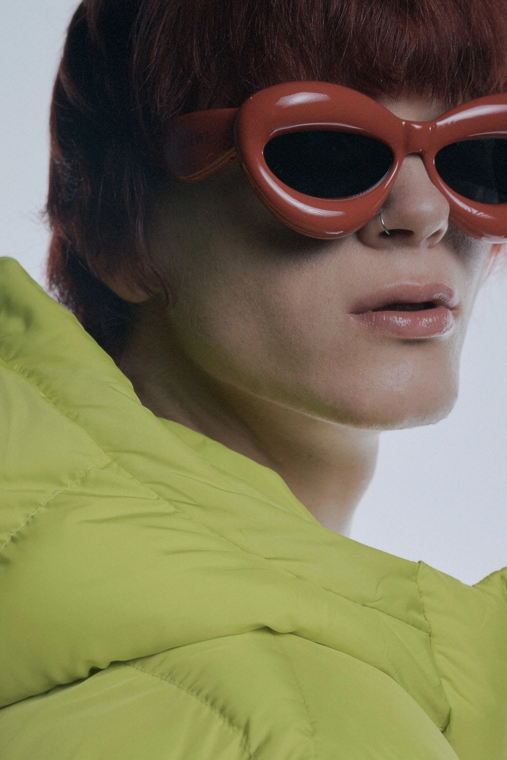 Nahaufnahme eines Menschen mit roten Haaren, trägt große, rote, schmale Sonnenbrille und ein silbernes Nasenpiercing. Er trägt einen leuchtend gelben Jacke.