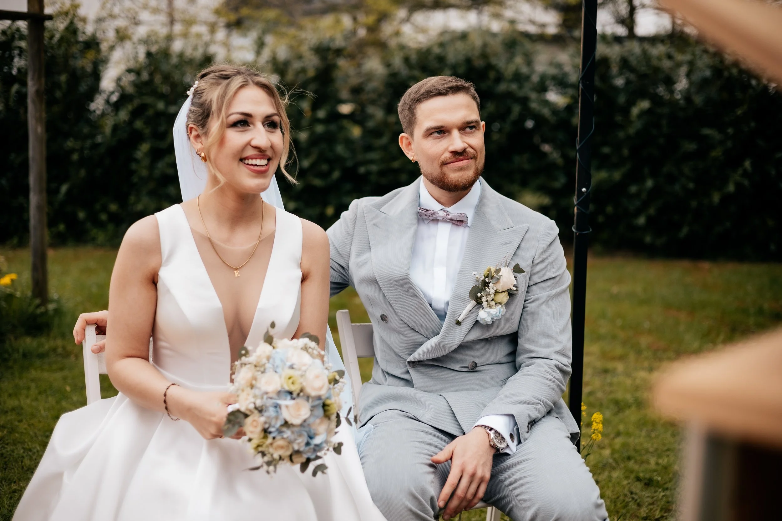 Ein glückliches Paar bei einer Hochzeit im Freien, die Braut trägt ein weißes Kleid und hält einen Blumenstrauß, der Bräutigam trägt einen hellgrauen Anzug mit Fliege.