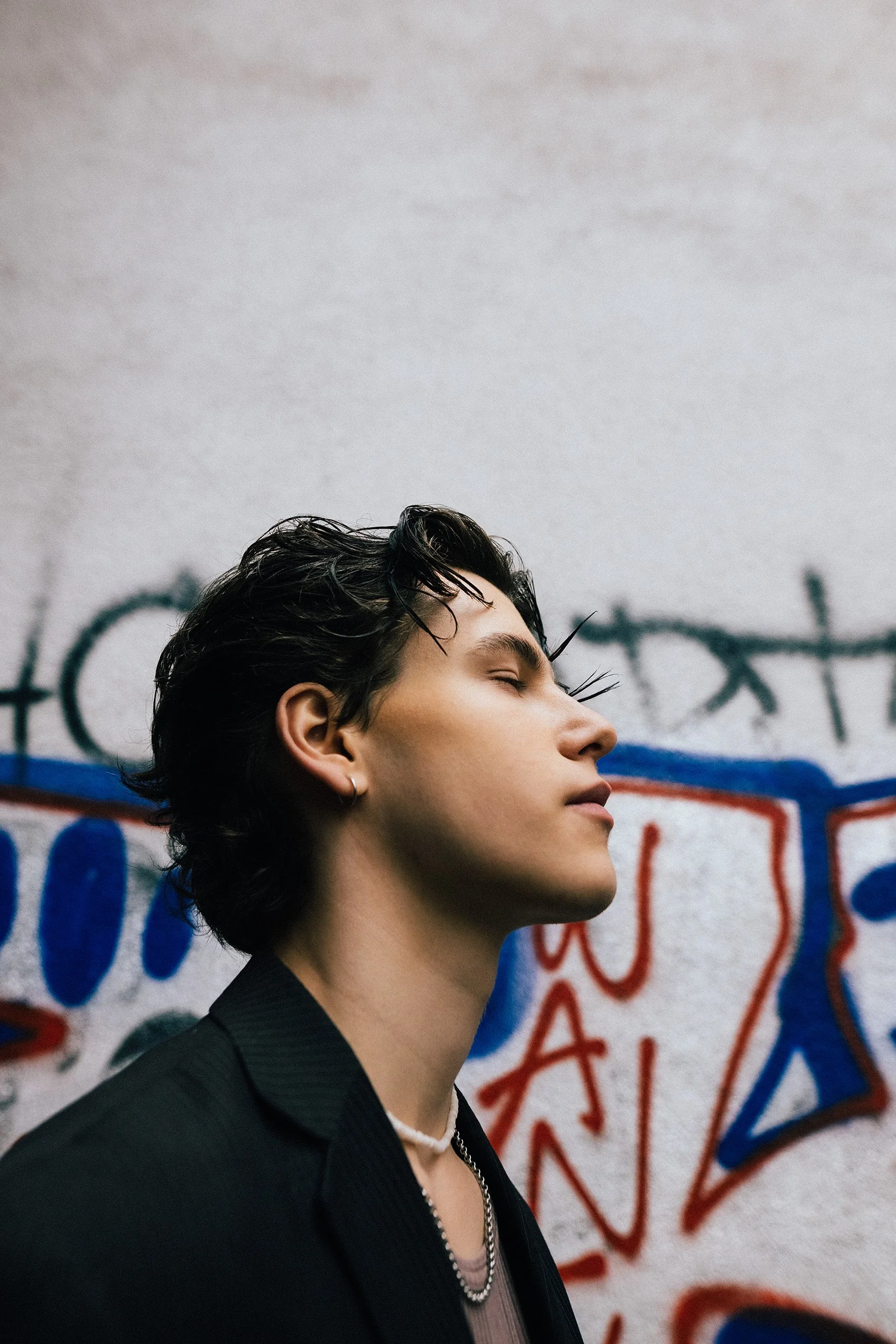 Porträt eines jungen Mannes mit kurzen, nassen dunklen Haaren, in schwarzer Jacke und mit Kette, vor einer Graffiti-Wand
