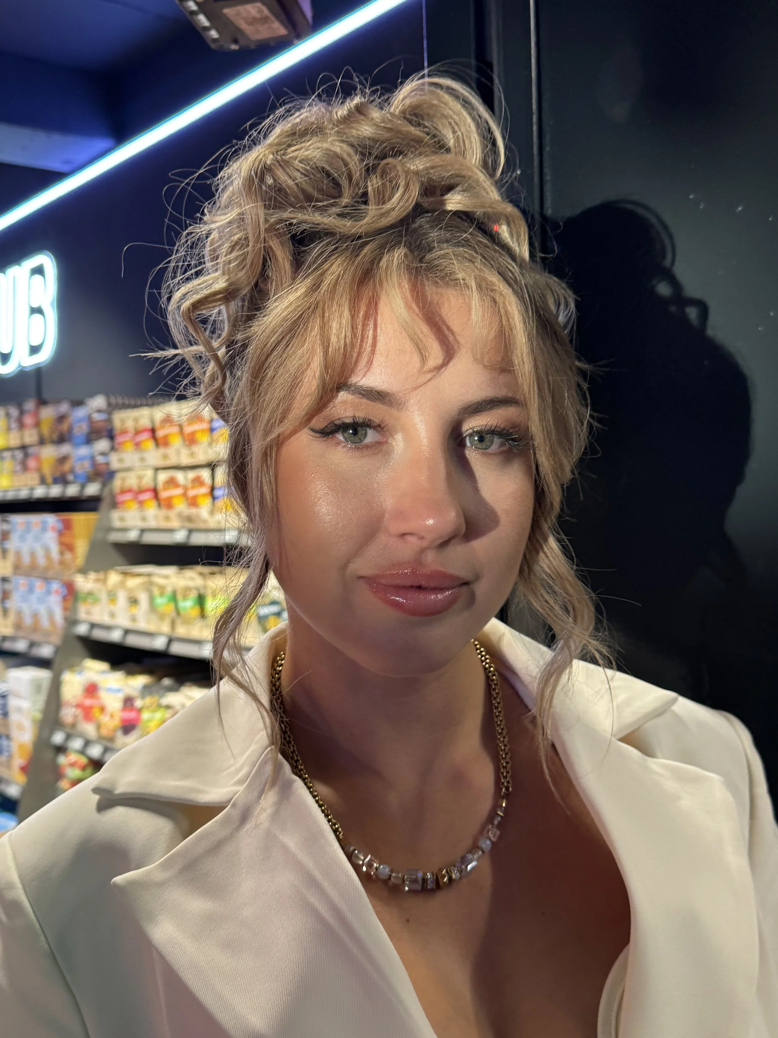 Nahaufnahme eines jungen Mädchens mit lockigem, blondem Haar. Sie trägt einen weißen Blazer und eine Perlenkette. Im Hintergrund sind Regale mit Snacks und Süßigkeiten in einem Supermarkt sichtbar. Die Beleuchtung ist warm, und es scheint nachts oder