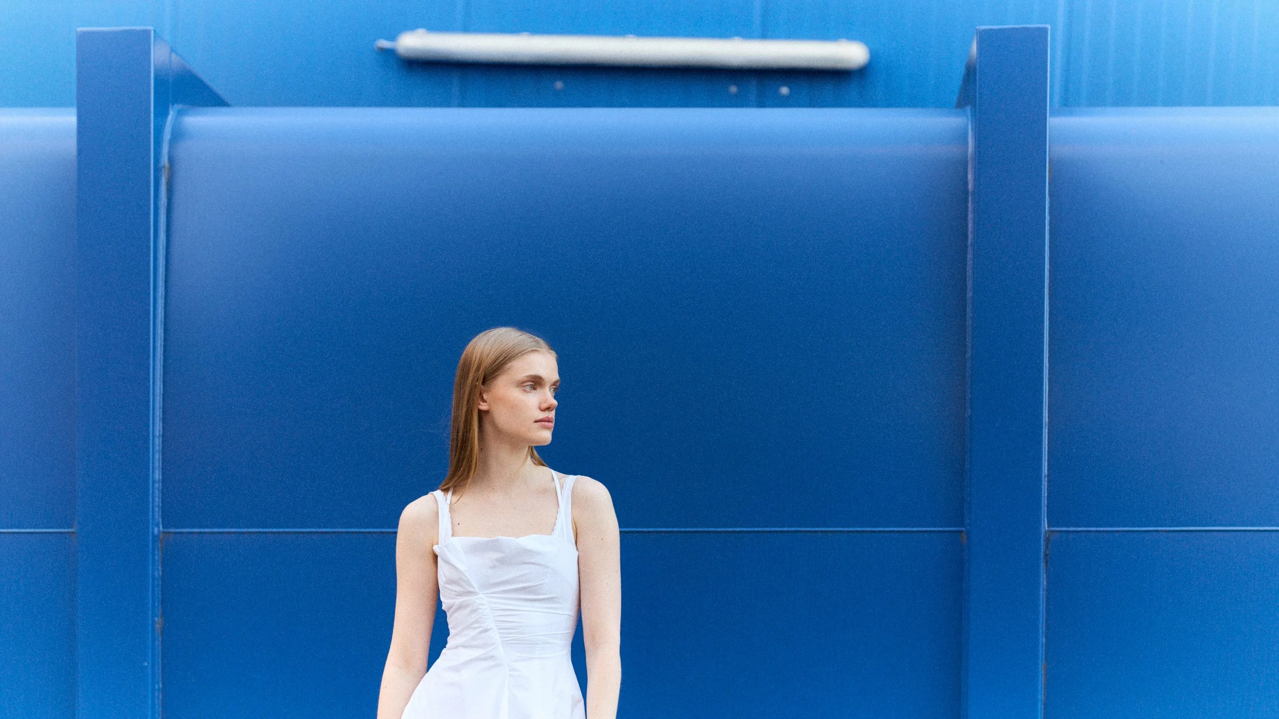 Junge Frau mit weißen Kleid vor blauer Wand, seitlich fotografiert.