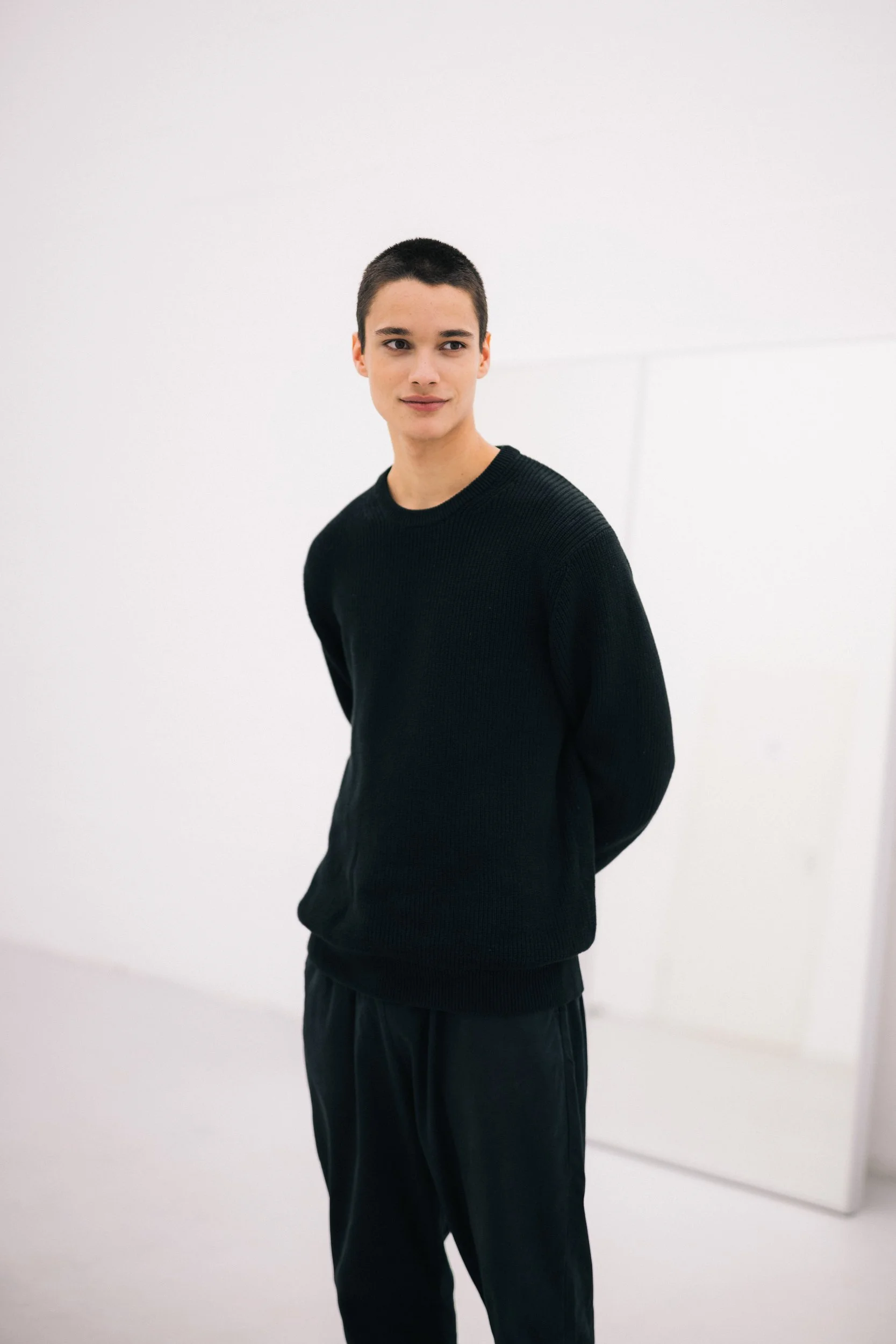 Junge Person mit kurzen dunklen Haaren, trägt schwarzen Pullover und schwarze Hosen, steht vor weißer Wand, blickt nach rechts, lächelt leicht.
