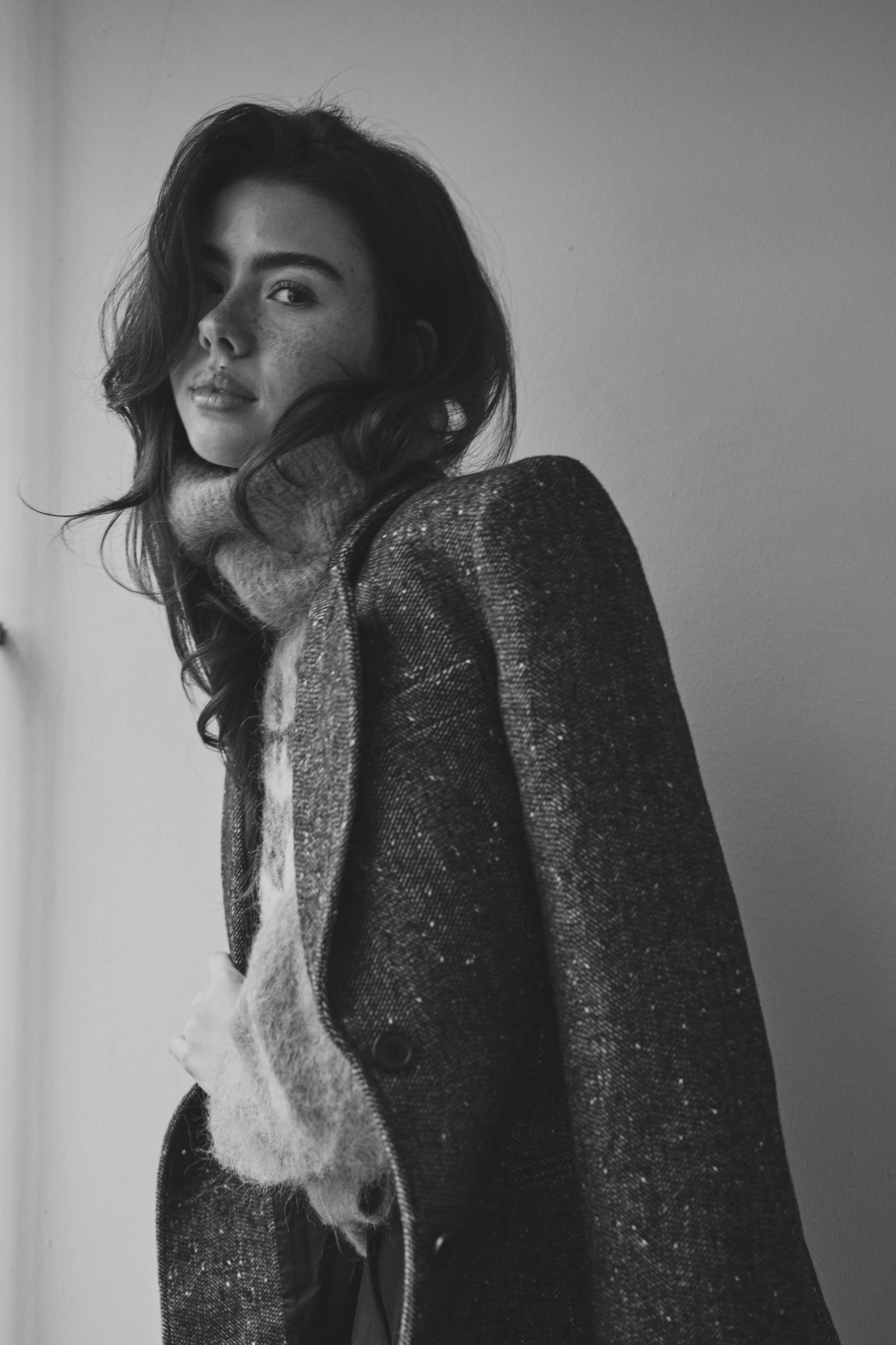 Schwarze und weiße Portraitaufnahme einer jungen Frau mit Locken, die eine Strickjacke trägt, vor einer schlichten Wand. Sie schaut direkt in die Kamera, mit einem leichten Lächeln.