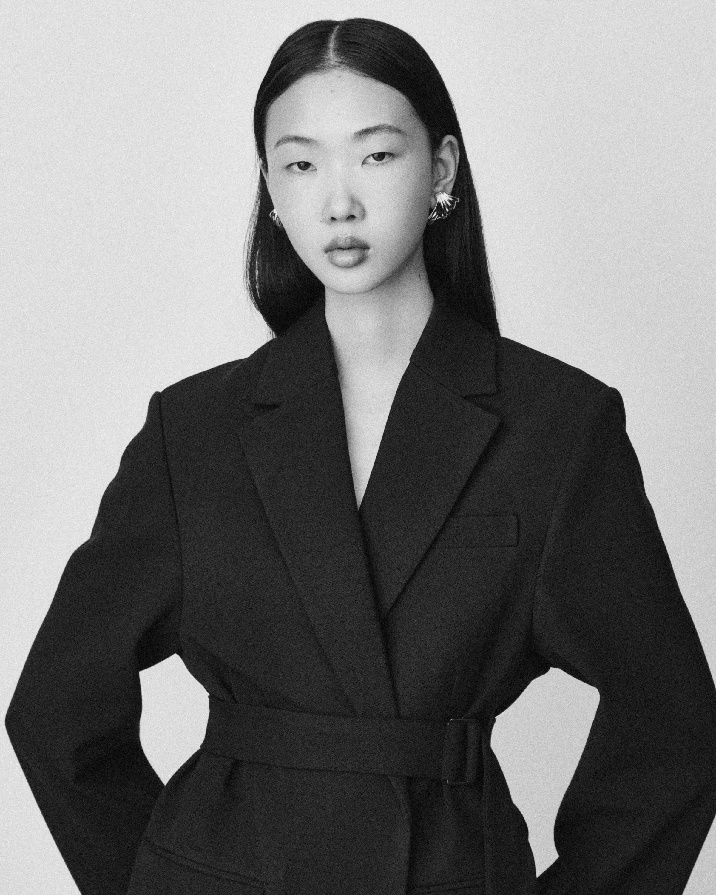 Schwarz-weiße Fotografie einer jungen asiatischen Frau mit langen, glatten Haaren. Sie trägt eine schwarze Blazerjacke mit einem Gürtel und auffällige Ohrringe. Der Hintergrund ist schlicht weiß.