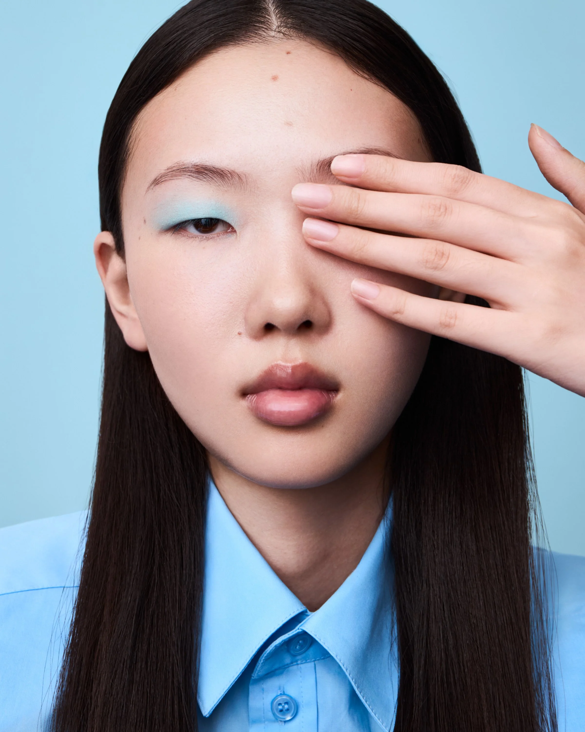 Nahaufnahme eines jungen Mädchens mit glatter Haut, braunen Haaren und blauer Bluse, das die rechte Augenpartie mit der Hand bedeckt, vor einem hellblauen Hintergrund.