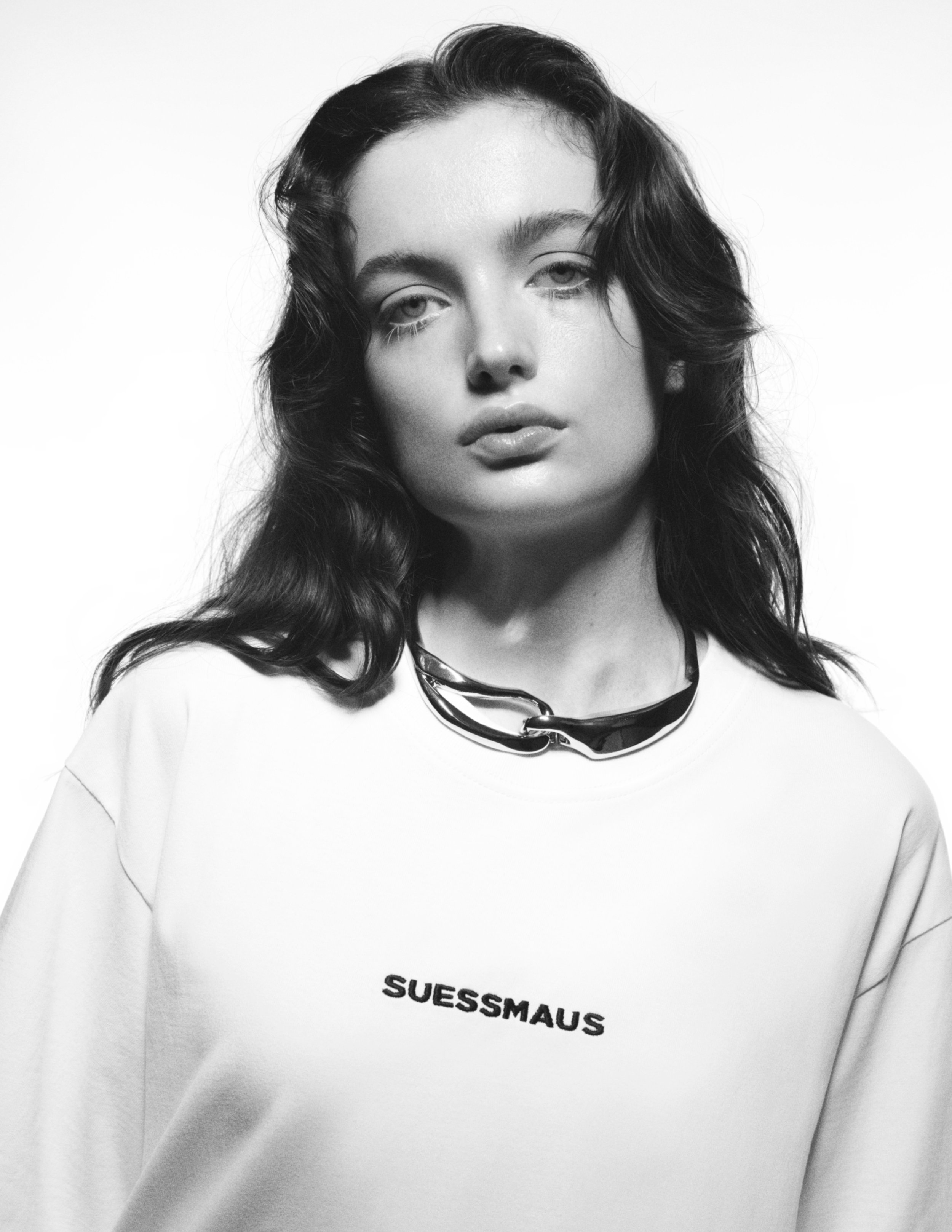 Schwarzweißfoto einer jungen Frau mit dunklen, gewellten Haaren, die ein T-Shirt mit dem Schriftzug »SUESSMALUS« trägt und eine große Kunststoffkette um den Hals hat.