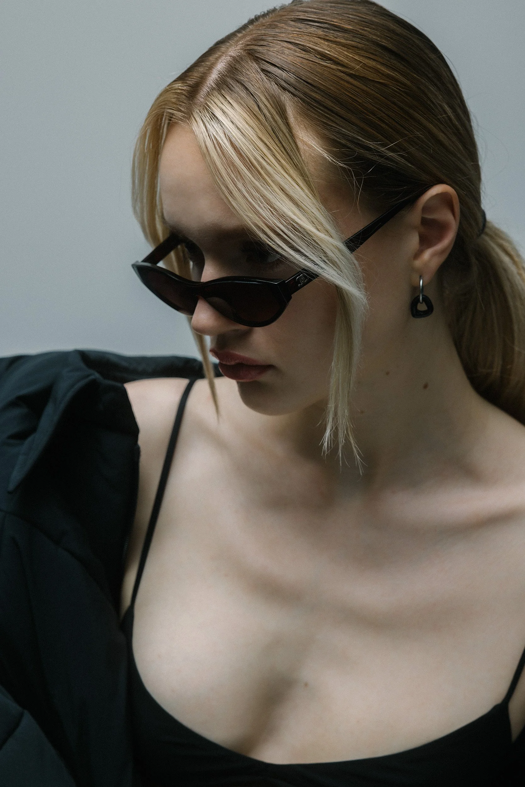Nahaufnahme eines jungen Frau mit blonden Haaren, Sonnenbrille und Piercing am Ohr, trägt ein schwarzes Oberteil.