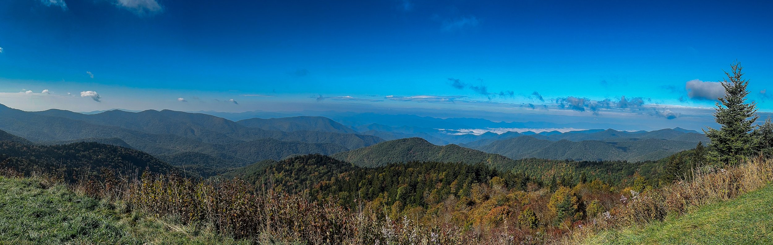 Panoramic view from the Blue Ridge Parkway overlooking layers of the Appalachian Mountains beneath a vivid blue sky on an autumn day.