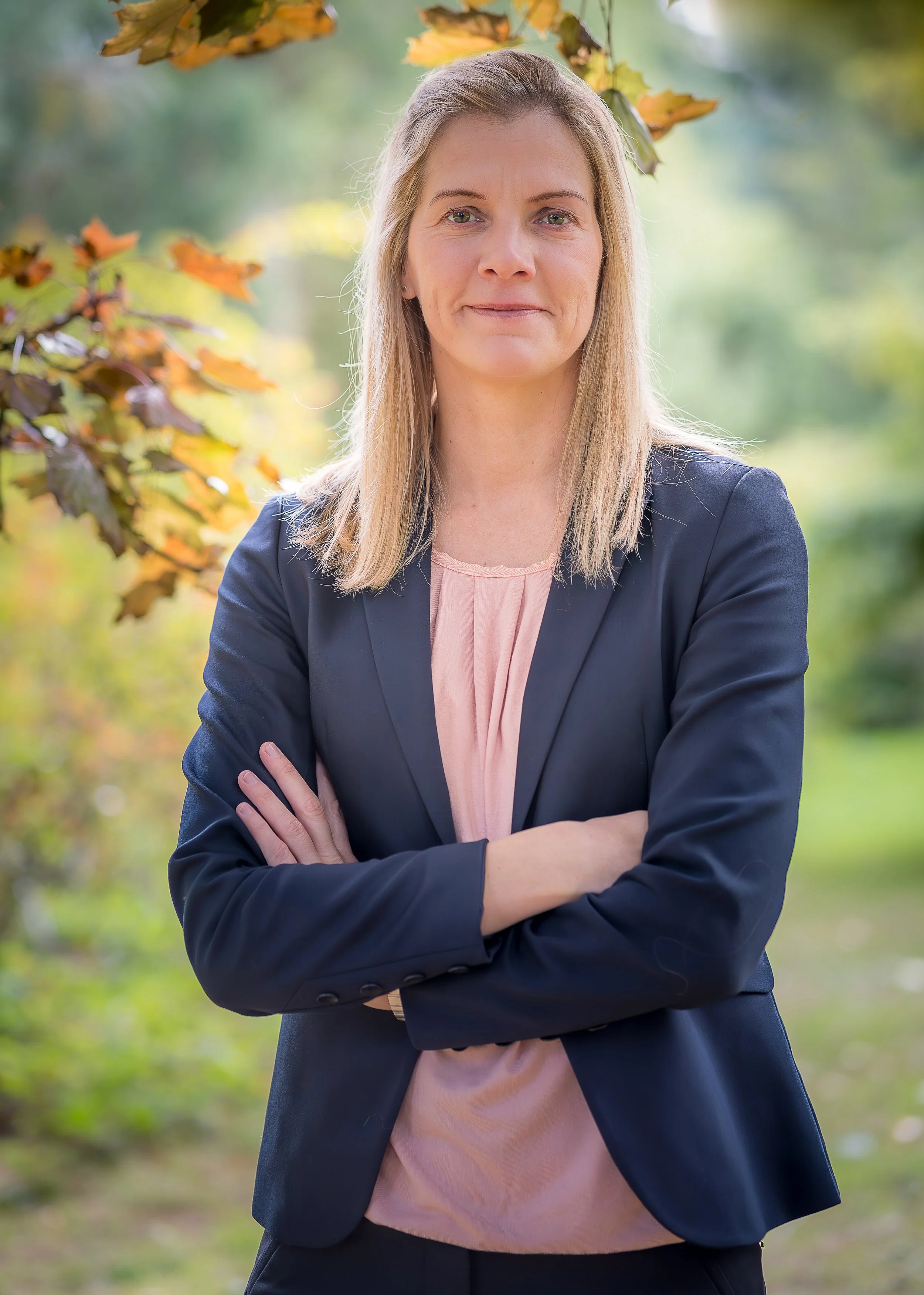 En portrætfotografi af en kvinde med skulderlangt blond hår, iført mørkeblåt jakkesæt og lyserødt top, der står udenfor med arme krydset over brystet, omgivet af efterårsblade og en grøn, sløret baggrund.