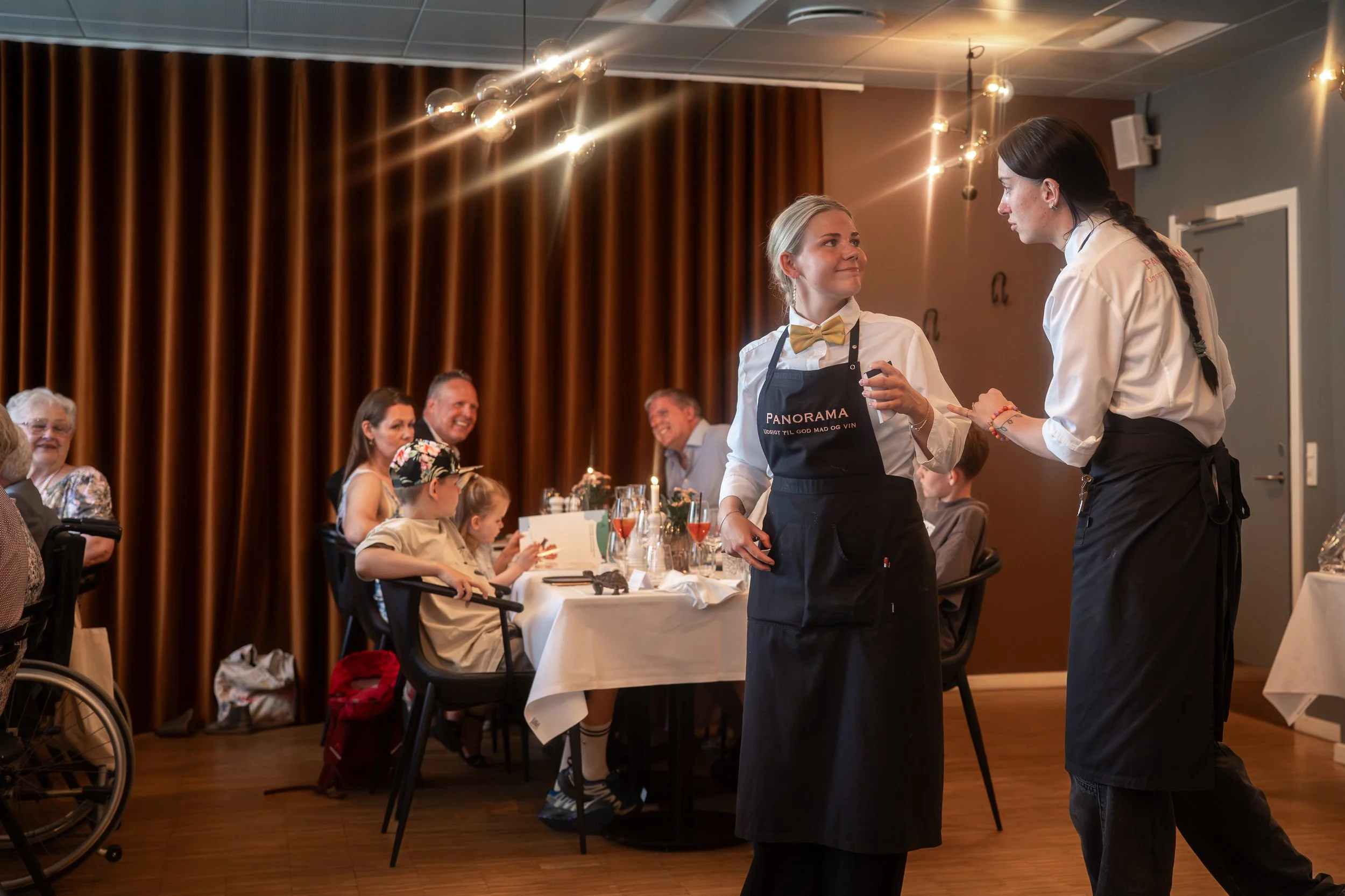 To tjenere smiler i forgrunden, mens gæsterne i restaurant Panorama hygger sig.