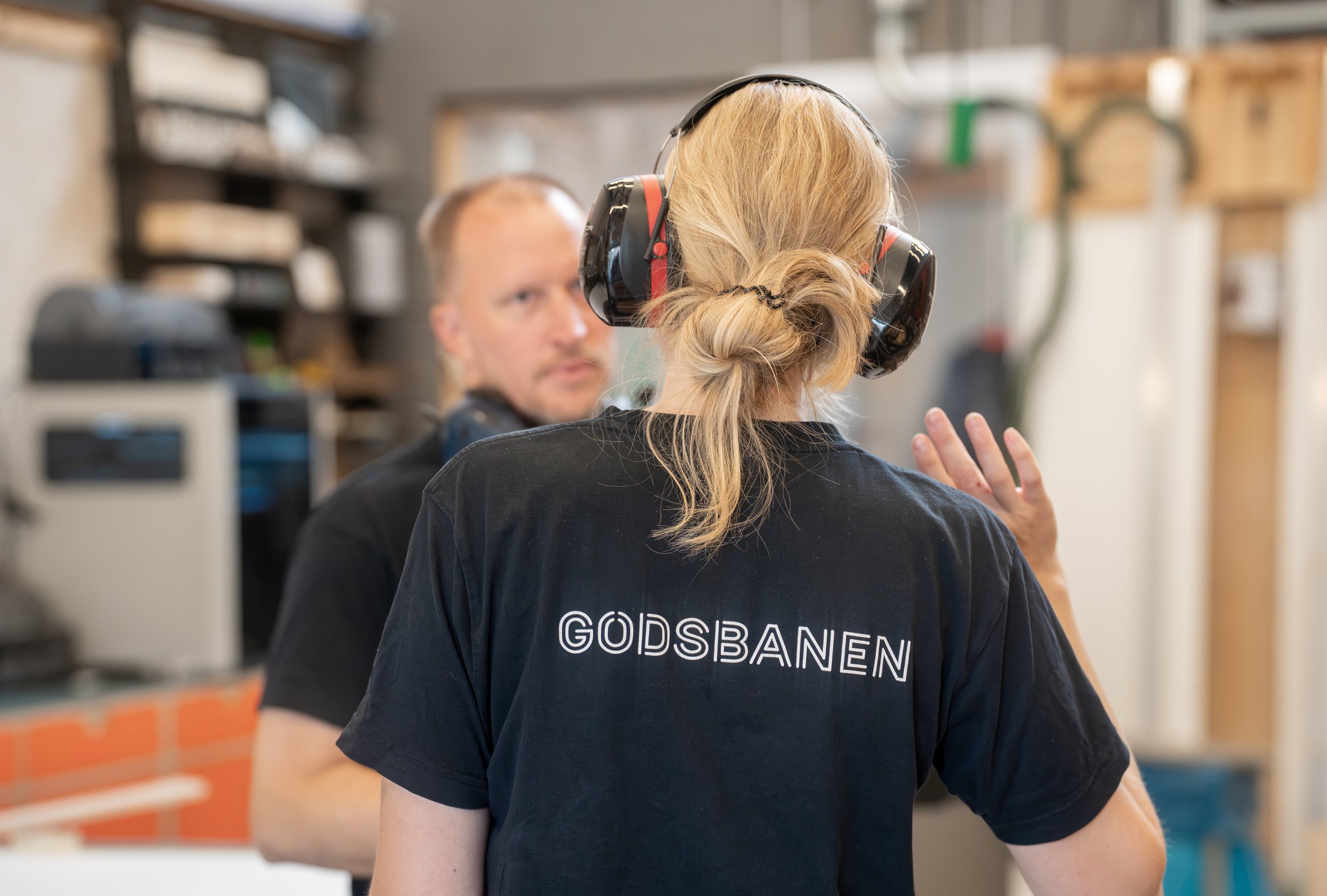 En kvinde med blond hår i hestehale, iført sort t-shirt med teksten 'GØDSBANEN', står foran en person med skulderlangt hår, begge iført høreværn, i en arbejds- eller værkstedsindstilling.