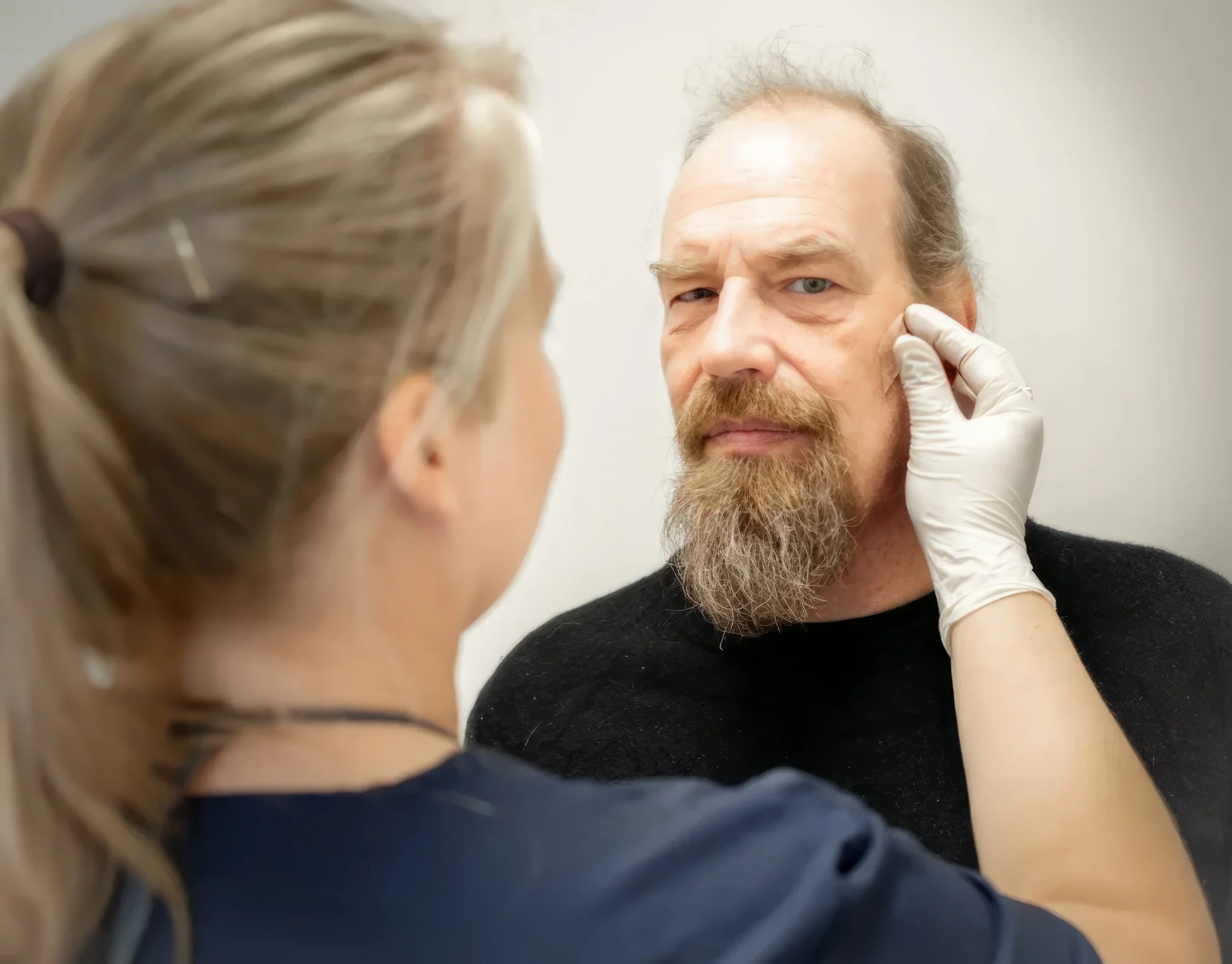 En mand med skæg, der bliver undersøgt af en person med latexhandsker, som tjekker mandens ansigt.