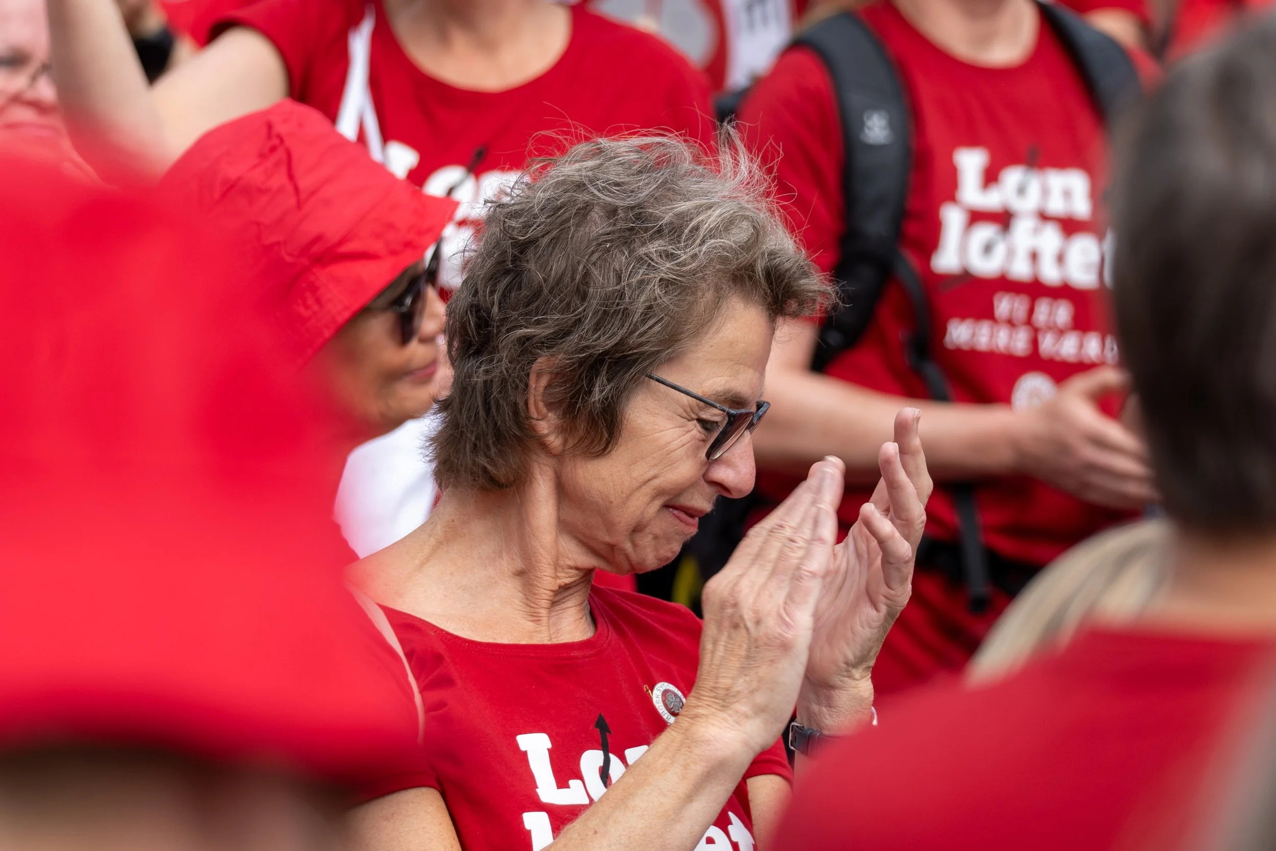 Gruppe mennesker til en demonstration iført røde T-shirts, nogle lytter opmærksomt og har lukkede øjne, ser ud til at være følelsesmæssigt engagerede.